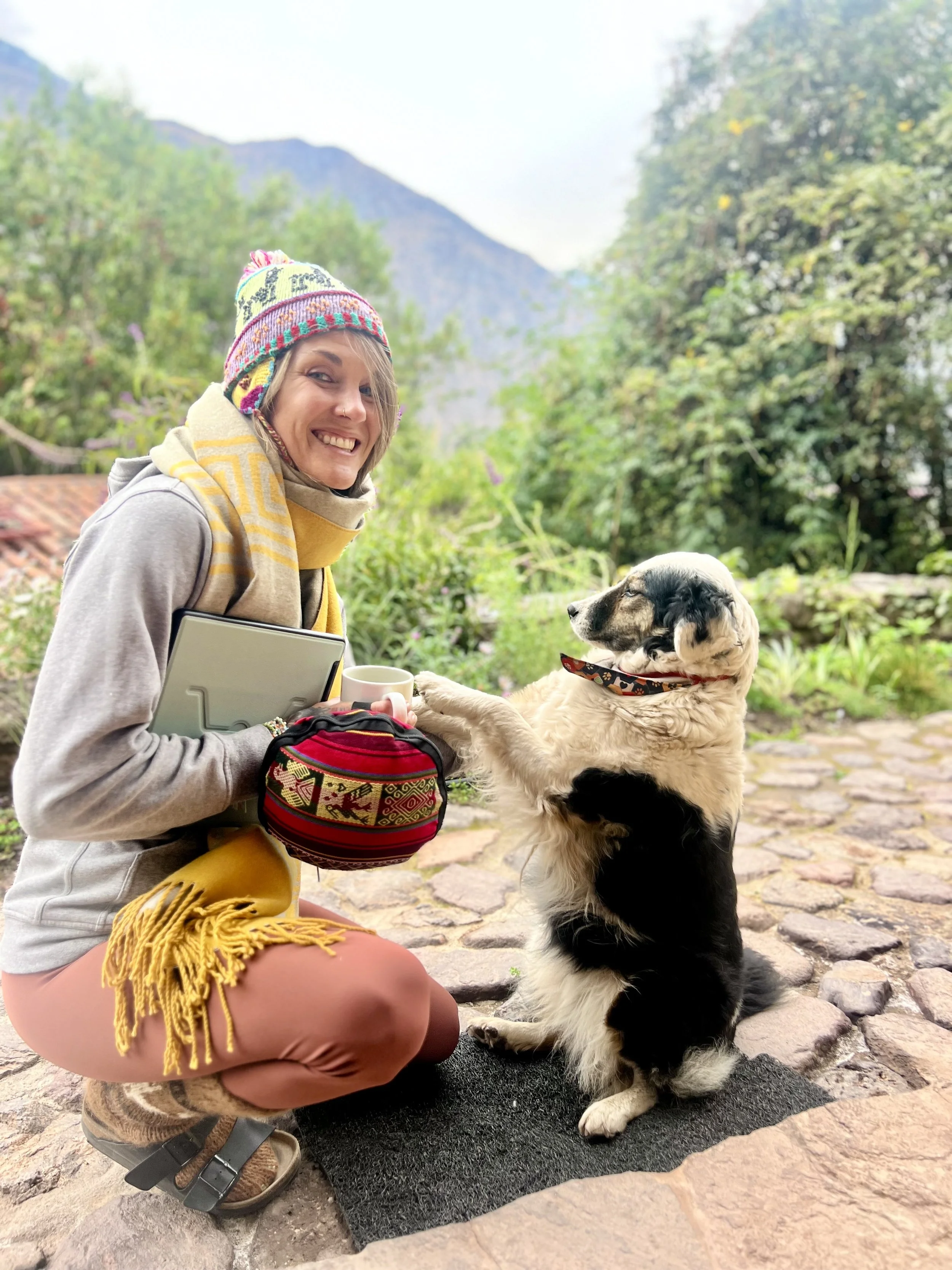 A woman smiling and holding a mug with her dog sitting on a doormat outdoors in a green mountain landscape.