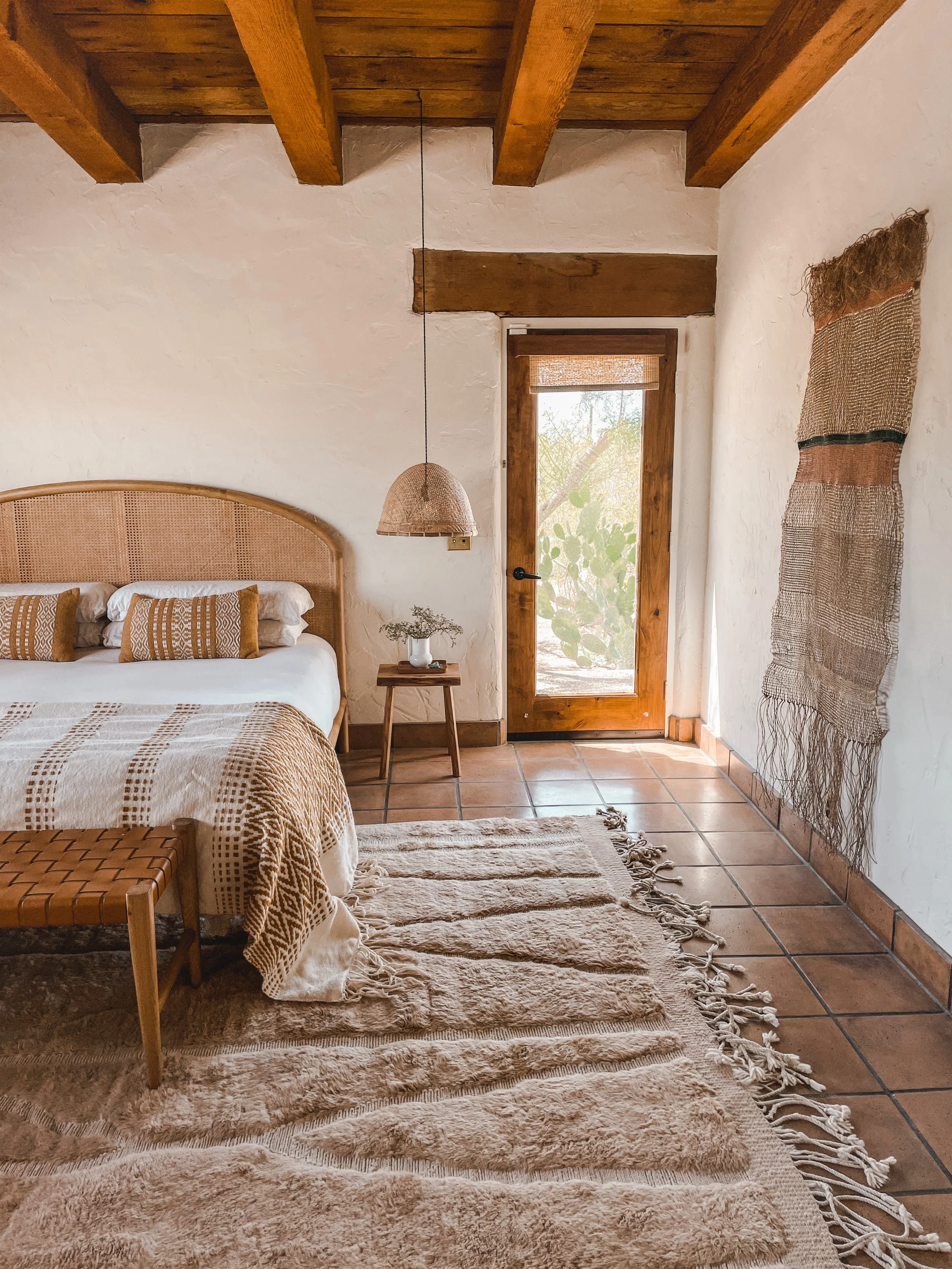 A cozy bedroom features earthy tones with a wooden ceiling and beams. There is a bed with a woven rattan headboard, decorated with patterned pillows and a bedspread. A plush beige rug covers the terracotta tiled floor. A small wooden side table holds a white vase with dried flowers, and a woven pendant lamp hangs from the ceiling. A door with a glass window leads outside, with greenery visible through it. A textured woven wall hanging decorates one white wall.