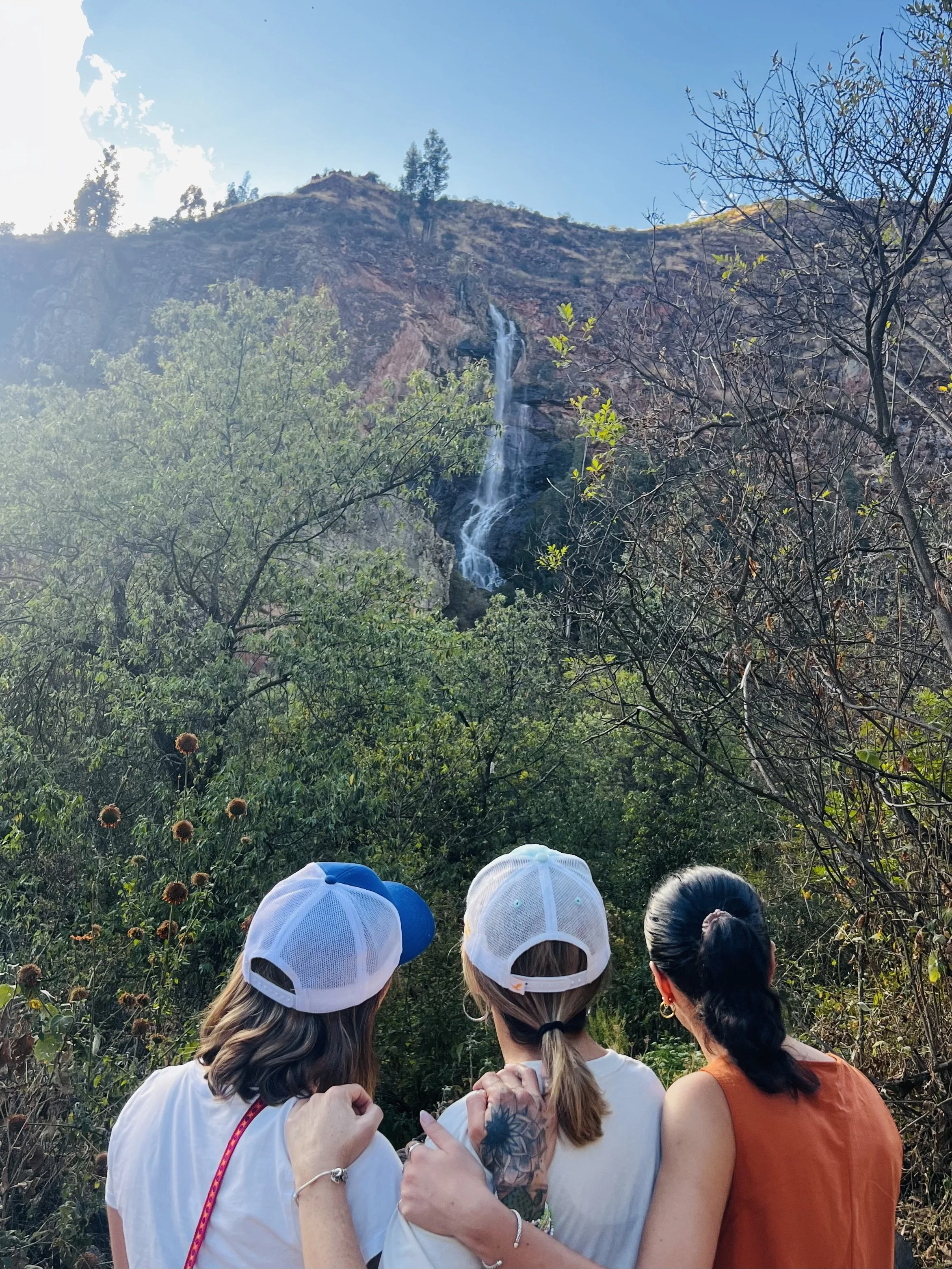 Three women in casual clothing and baseball caps look at a waterfall in a forested area with a mountain in the background.