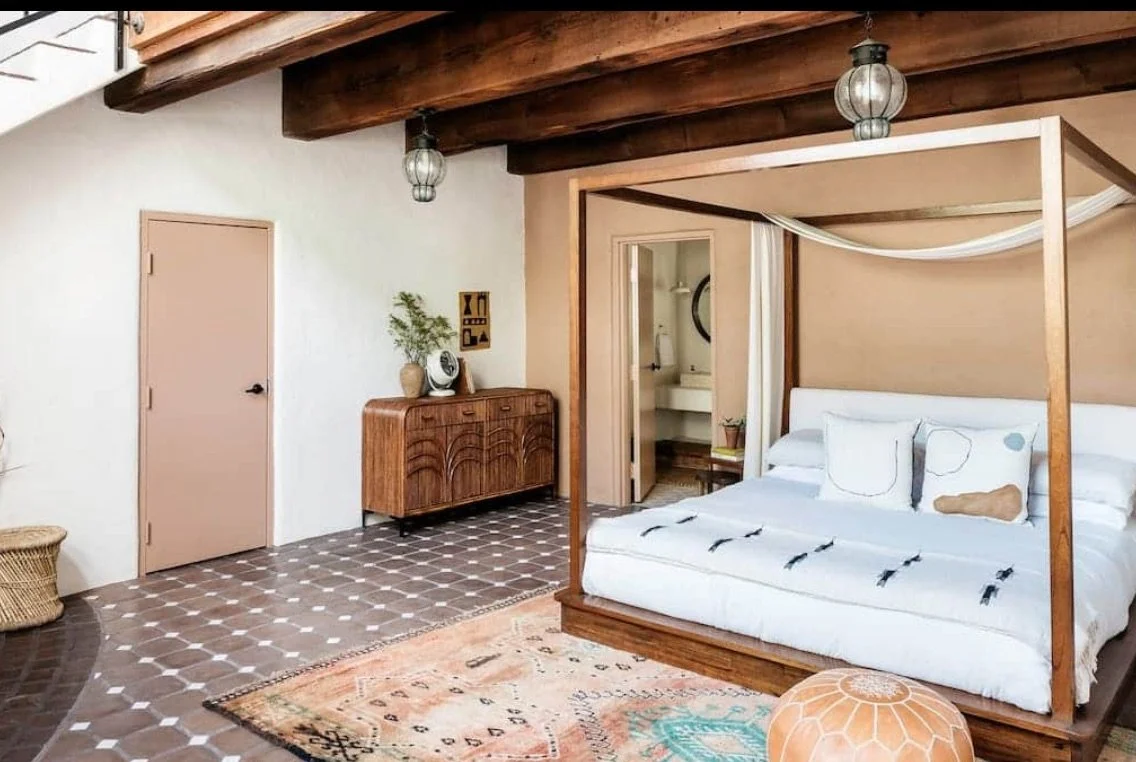 A bedroom with a four-poster bed, white bedding, and pillows, underneath wood beams with hanging lantern lights, a wicker basket, a wooden dresser with decorative items, and a patterned rug.