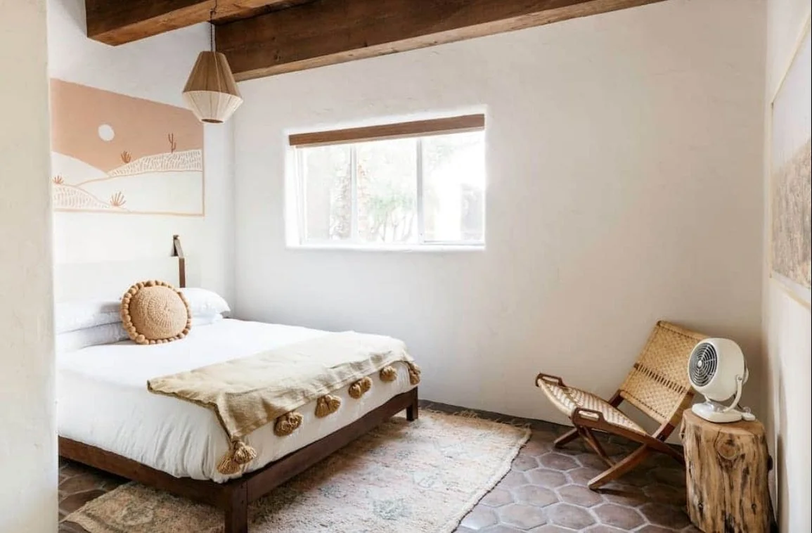 A cozy bedroom with white walls, wooden ceiling beams, a bed with white bedding and a decorative round pillow, a beige blanket with tassels, a landscape painting, a window with a wooden blind, a wooden chair, a small round wooden table with a fan, and a woven chair on a tiled floor.