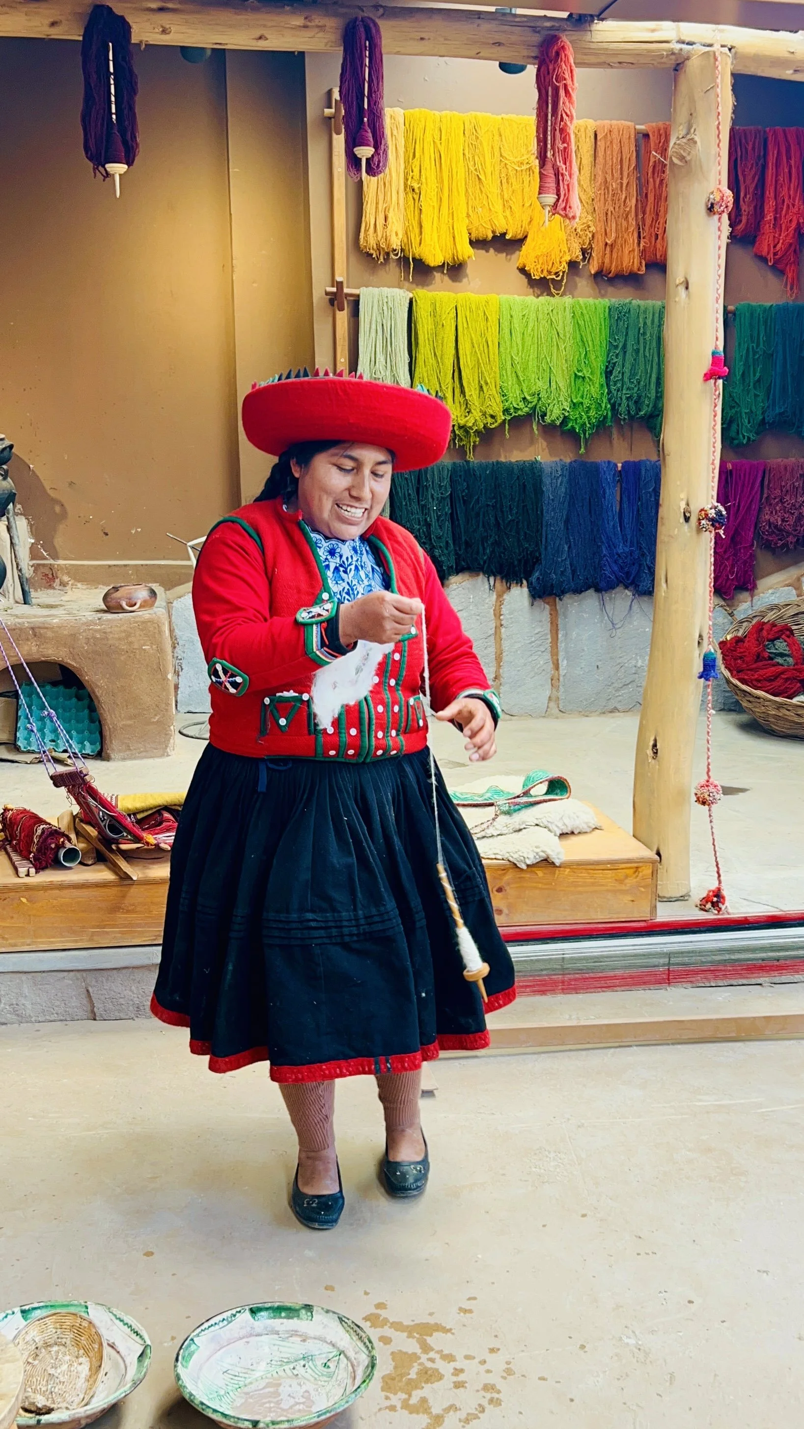 A woman in traditional Peruvian clothing, including a red hat and a black skirt, is knitting. Behind her are colorful yarns displayed on the wall, including yellow, green, blue, and purple. She is standing in a craft or textile shop or workshop.