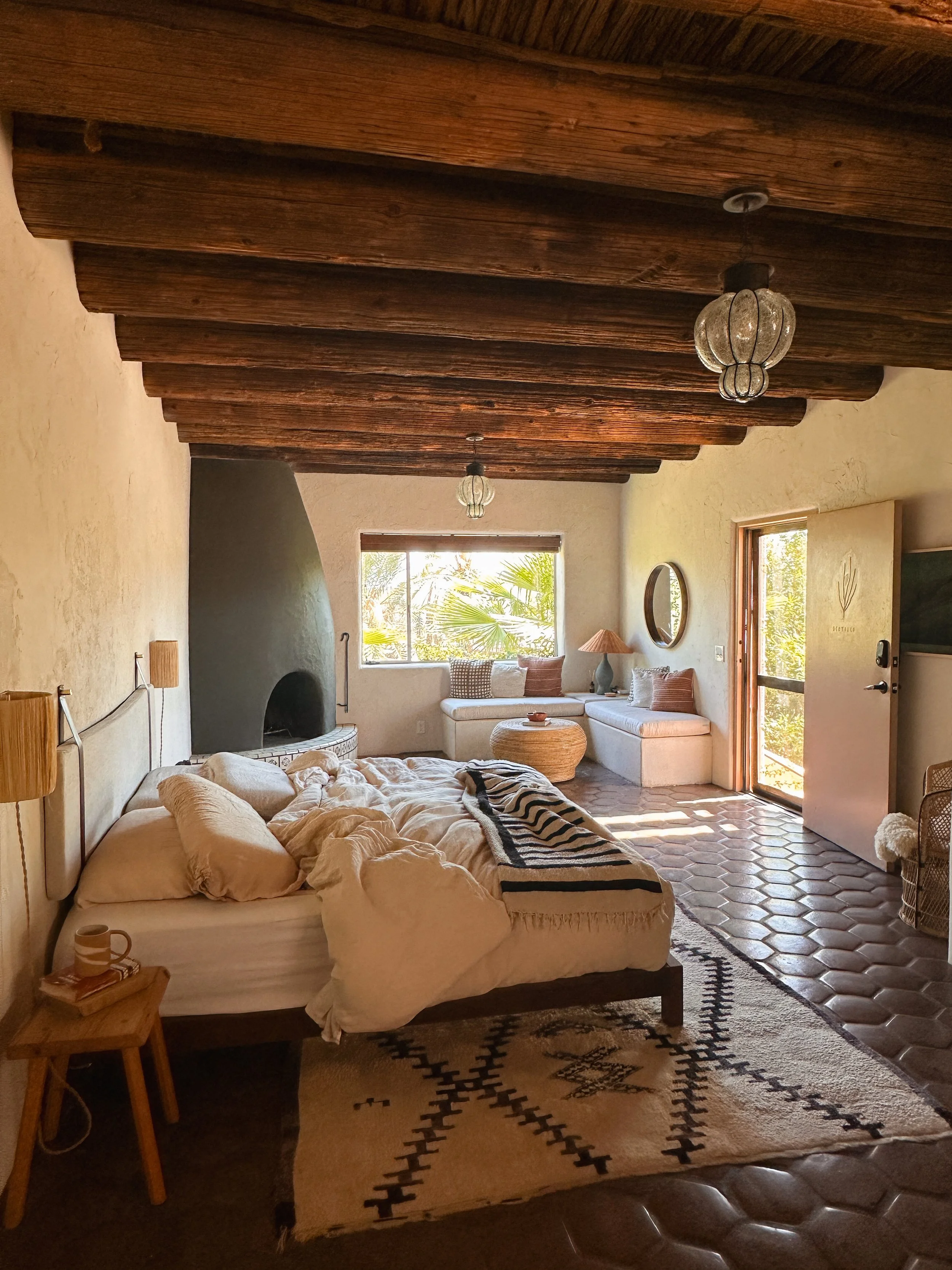 A cozy bedroom with a rustic wooden ceiling, a bed with cream-colored bedding, a small wooden side table with a mug, a patterned rug, a stone fireplace, a window with palm trees outside, a built-in corner couch with pillows, a round mirror, a lamp, and an open door leading outside.