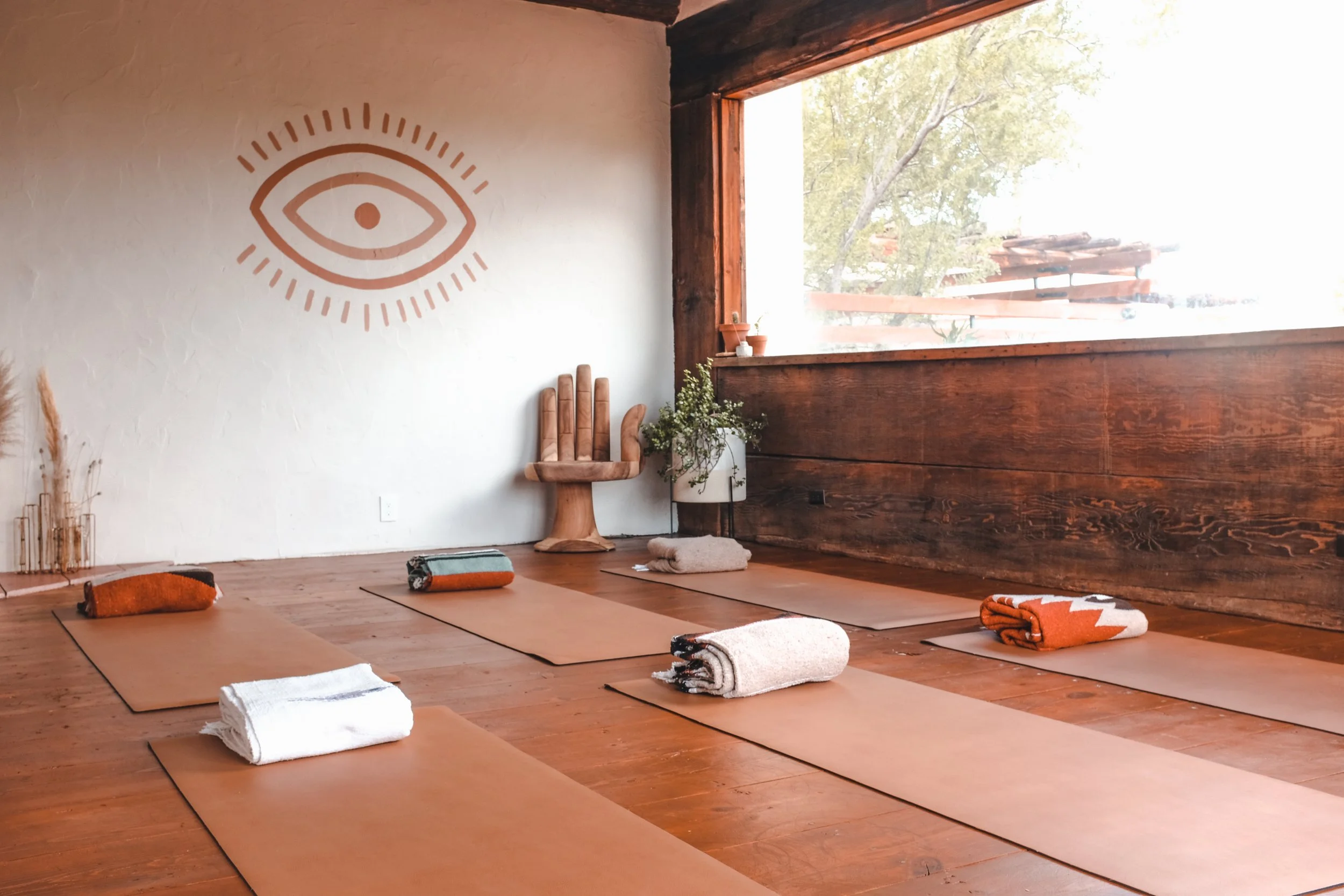 Yoga mats with rolled towels on a wooden floor in a peaceful studio with large window, decorative eye wall art, potted plant, and wooden chair.