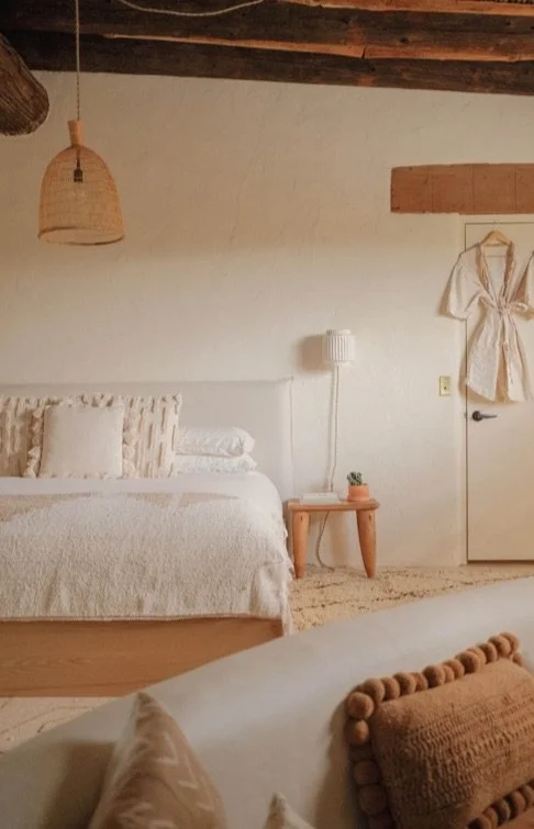 A cozy bedroom with white walls and wooden ceiling beams, a bed with neutral-colored linens, a small wooden side table with a plant, a hanging wicker light fixture, and a white dress hanging on the door.