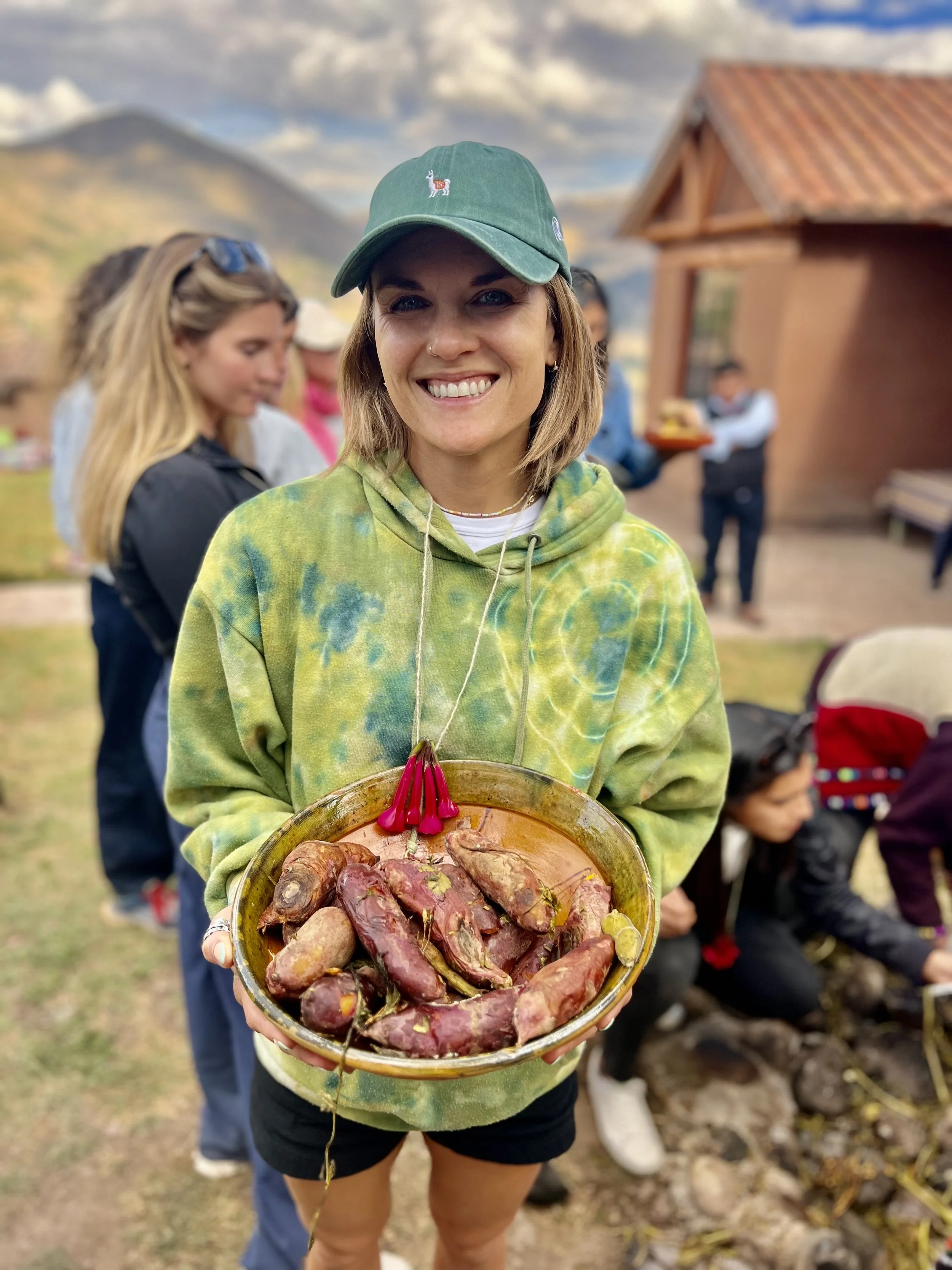 A woman smiling and holding a plate of cooked yams outdoors, with other people and a wooden building in the background.