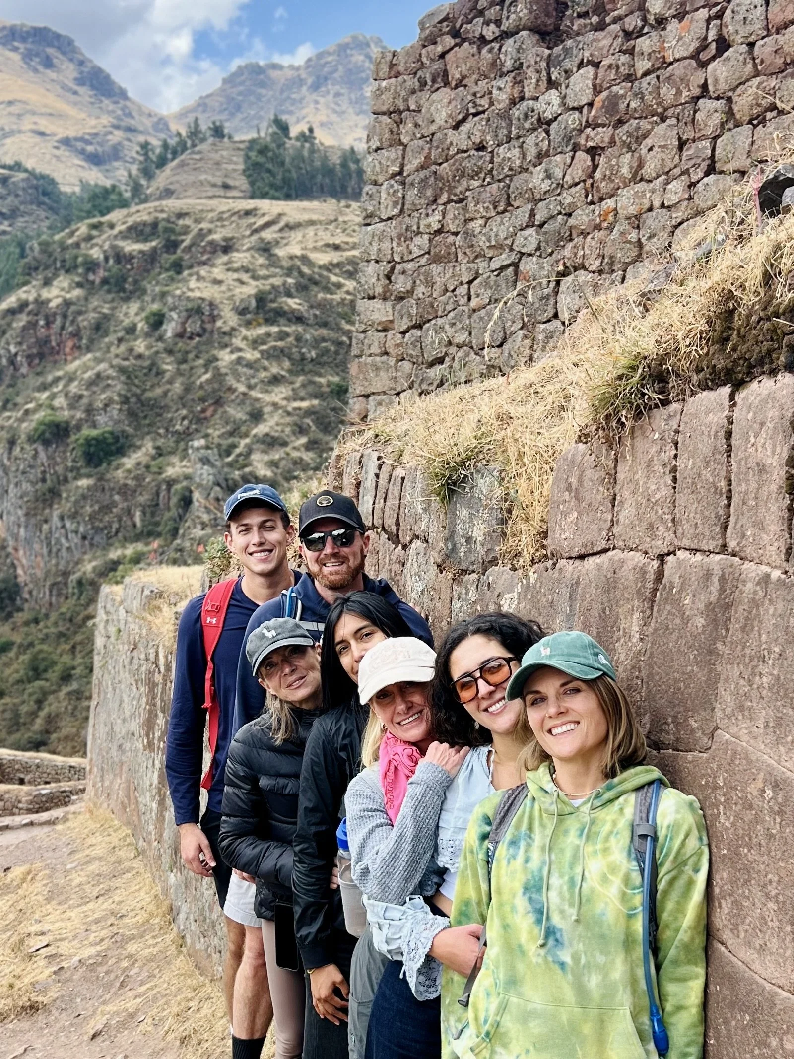 Group of six people outdoors beside an ancient stone wall, mountain landscape in the background, smiling for the camera.