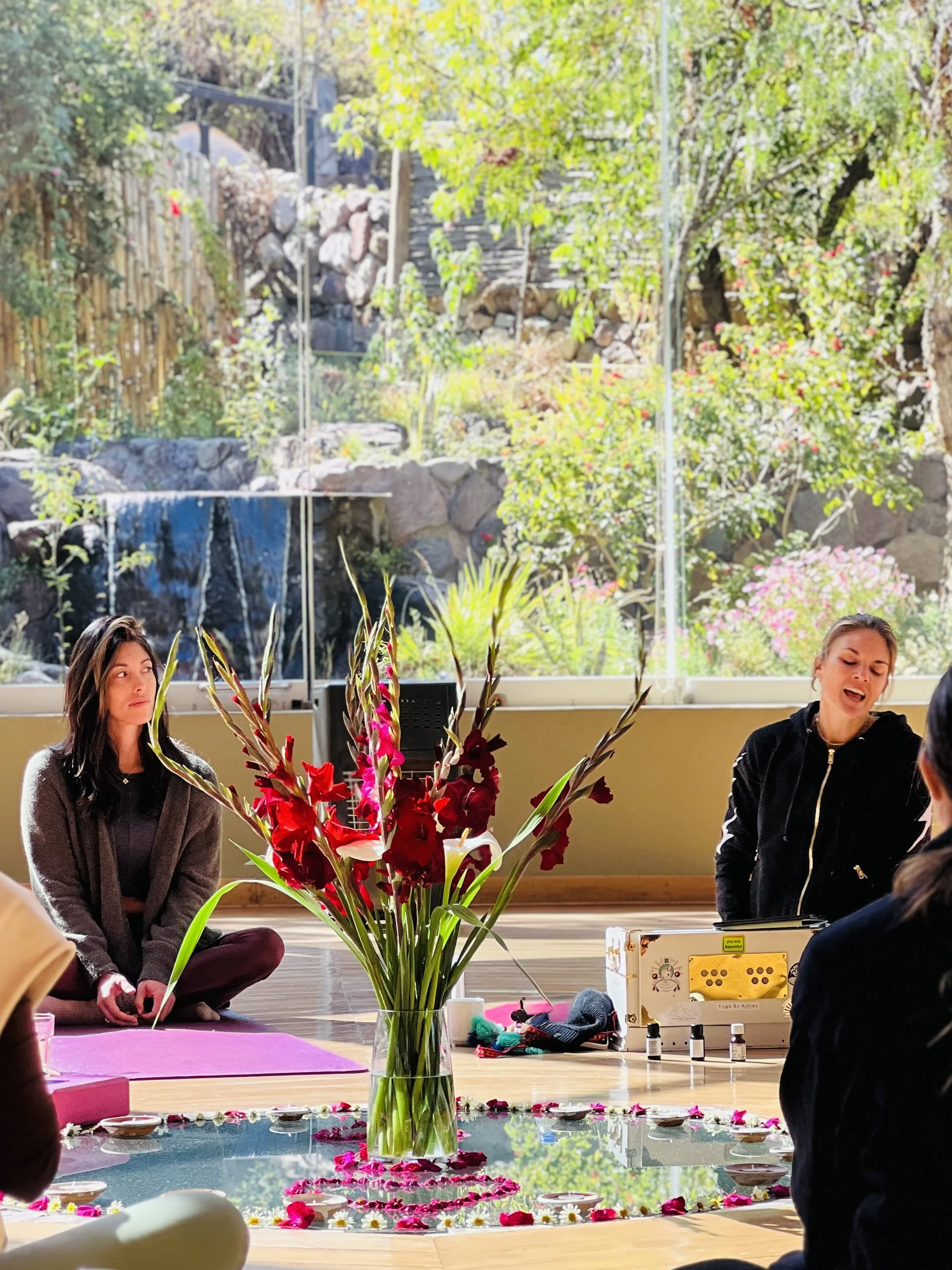 People participating in a meditation or spiritual session in a room with large windows overlooking a garden. There is a large glass vase with red and pink flowers on a mirror-like surface in the center of the room.