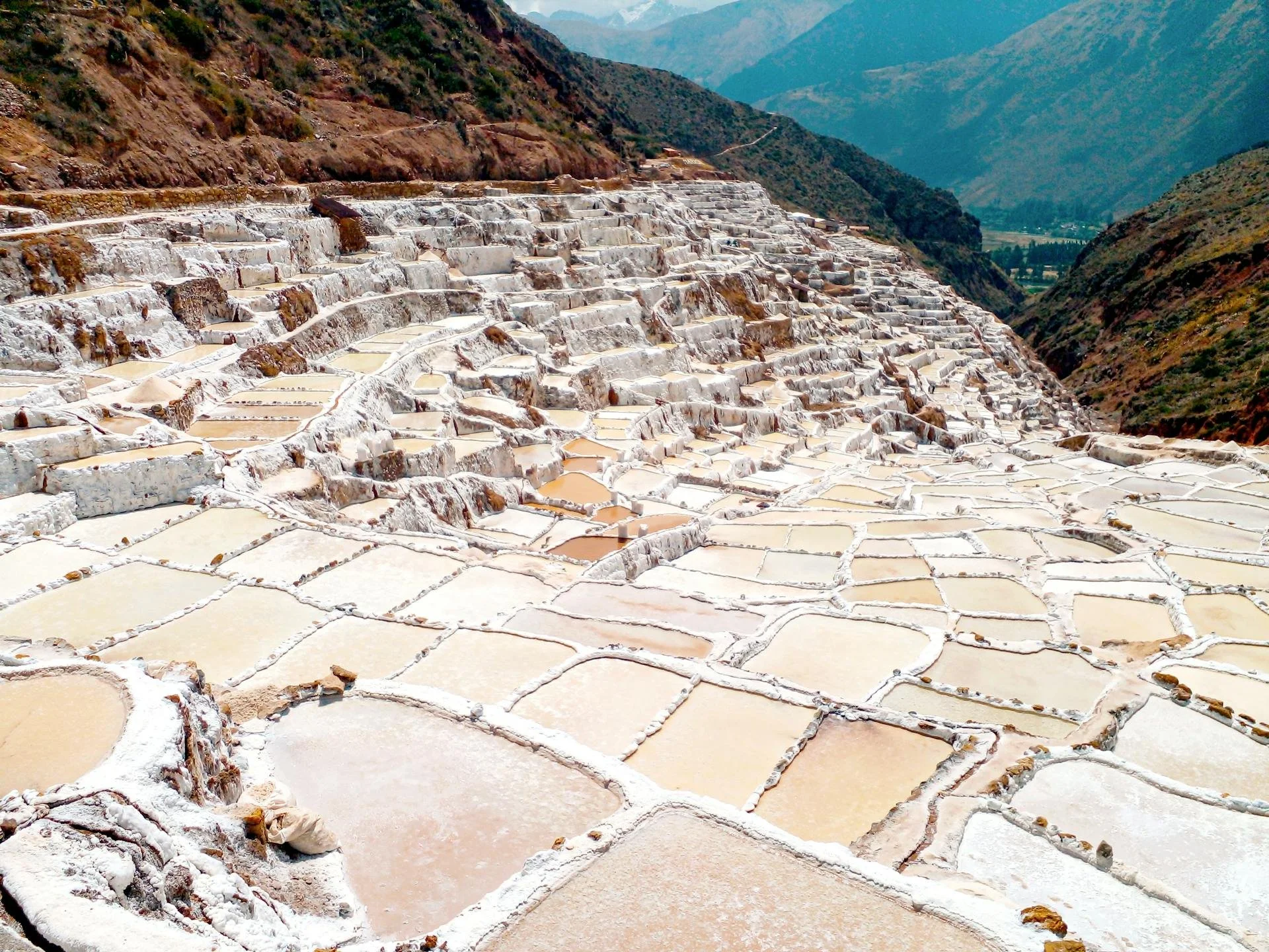 Panoramic view of terraced salt pans on a hillside, with mountains in the background. The salt pools form geometric shapes and vary in color from white to light brown.