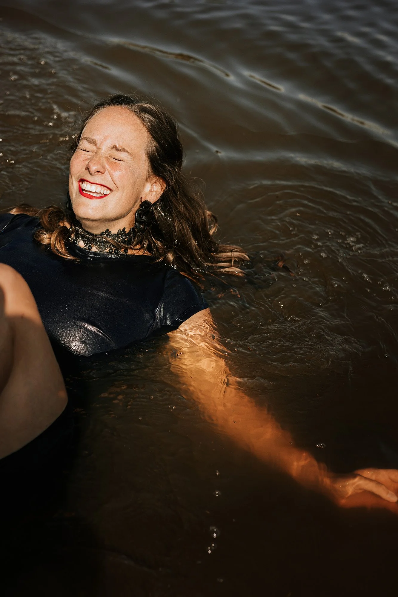 Vrouw met bruin haar, rode lippen en sieraden geniet van zwemmen in een rivier of meer, glimlachend met gesloten ogen.