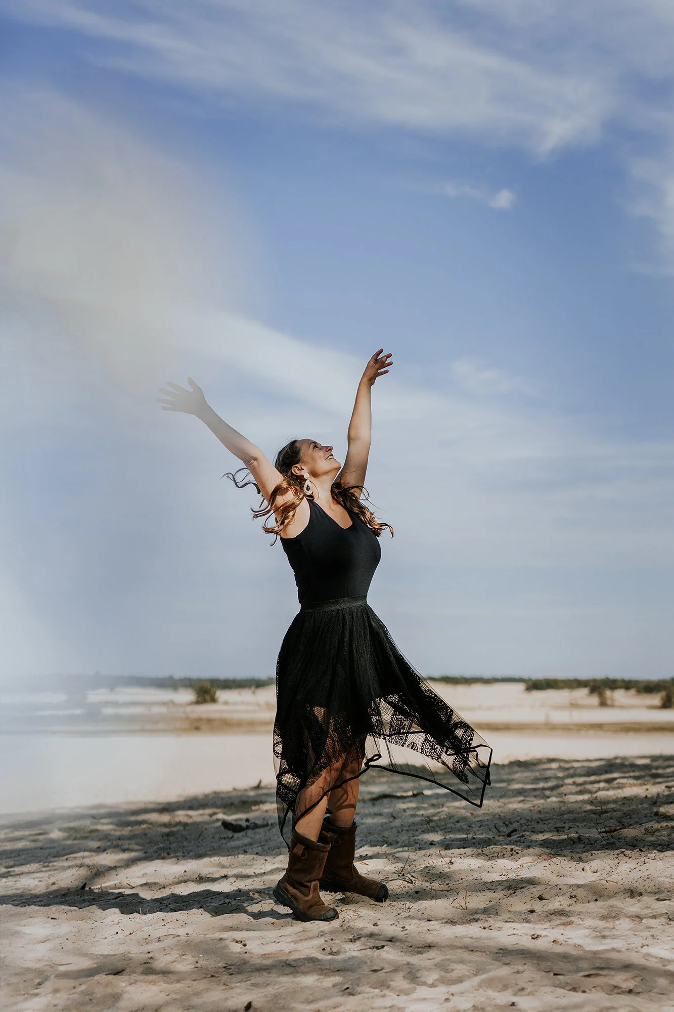 Vrouw in een zwart kanten rok en laarzen danst op het strand met haar armen omhoog, onder een blauwe lucht met wolken.