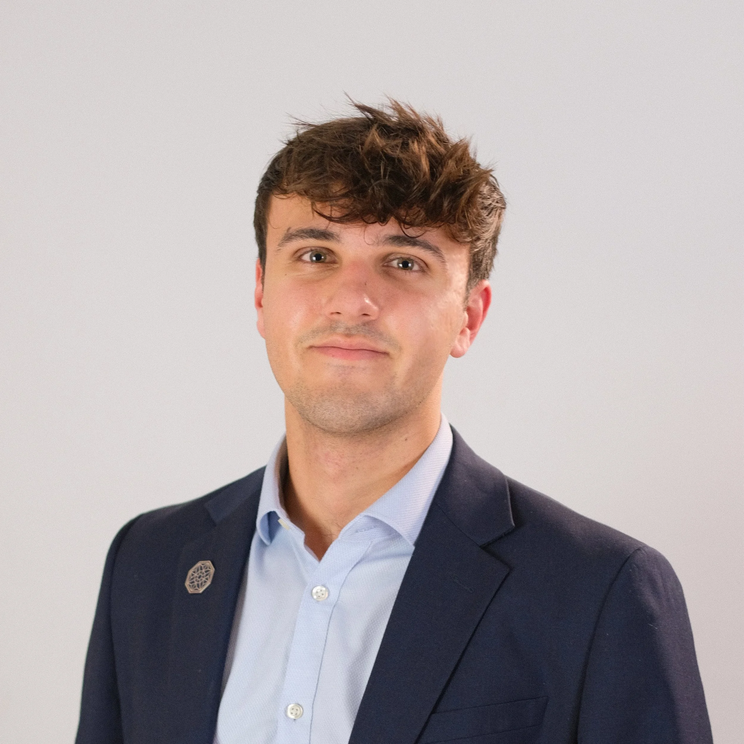 A young man with dark curly hair, wearing a light blue button-up shirt, standing outdoors in front of a large body of water and historic European-style buildings.