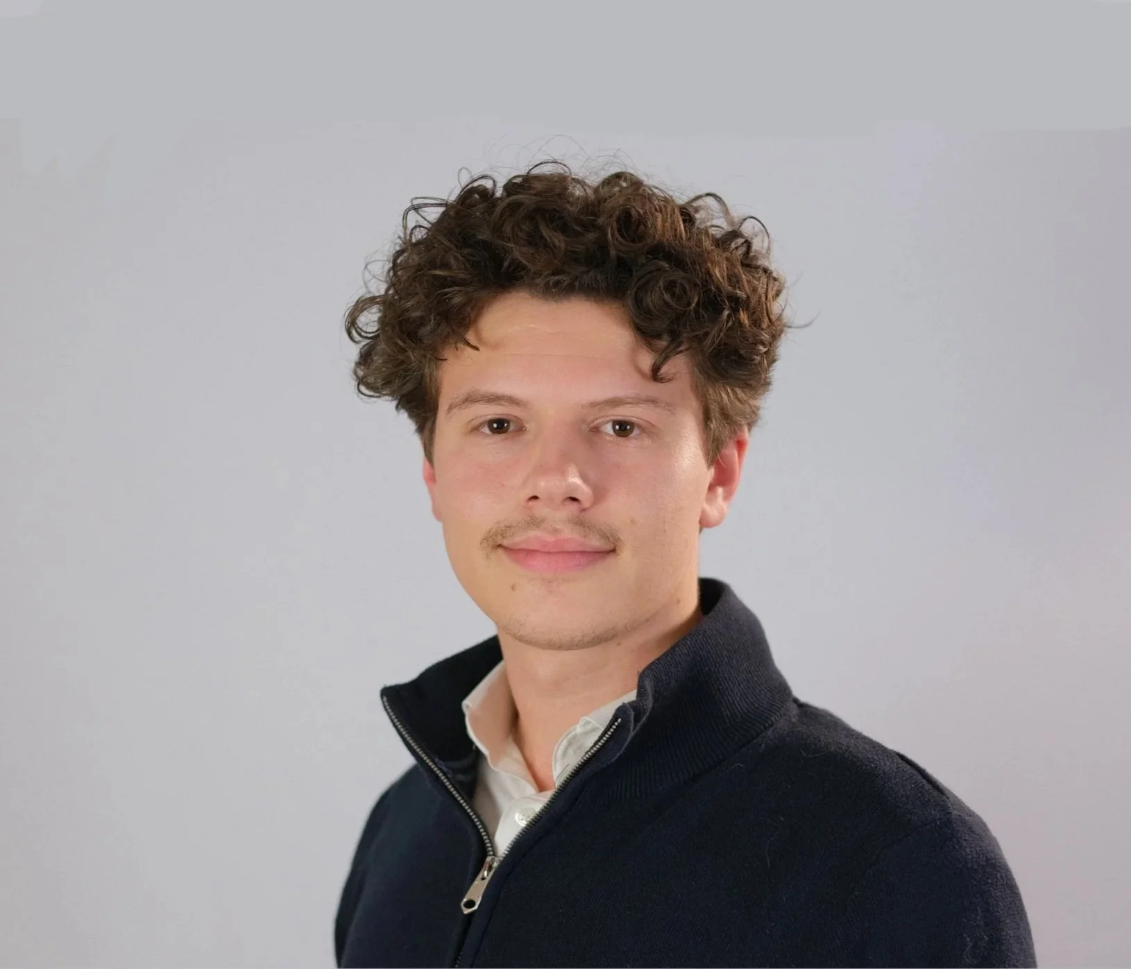 Young man with curly brown hair wearing a light blue checkered shirt, standing outdoors near water with buildings and trees in the background.