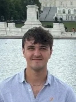 A young man with dark curly hair, wearing a light blue button-up shirt, standing outdoors in front of a large body of water and historic European-style buildings.