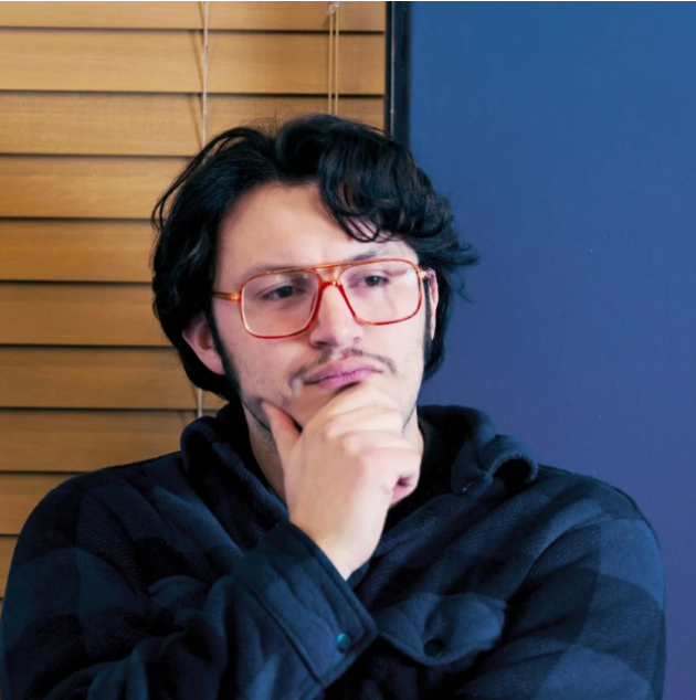 A young man with glasses holding a snake, looking at it with curiosity inside a room with blinds covering a window.