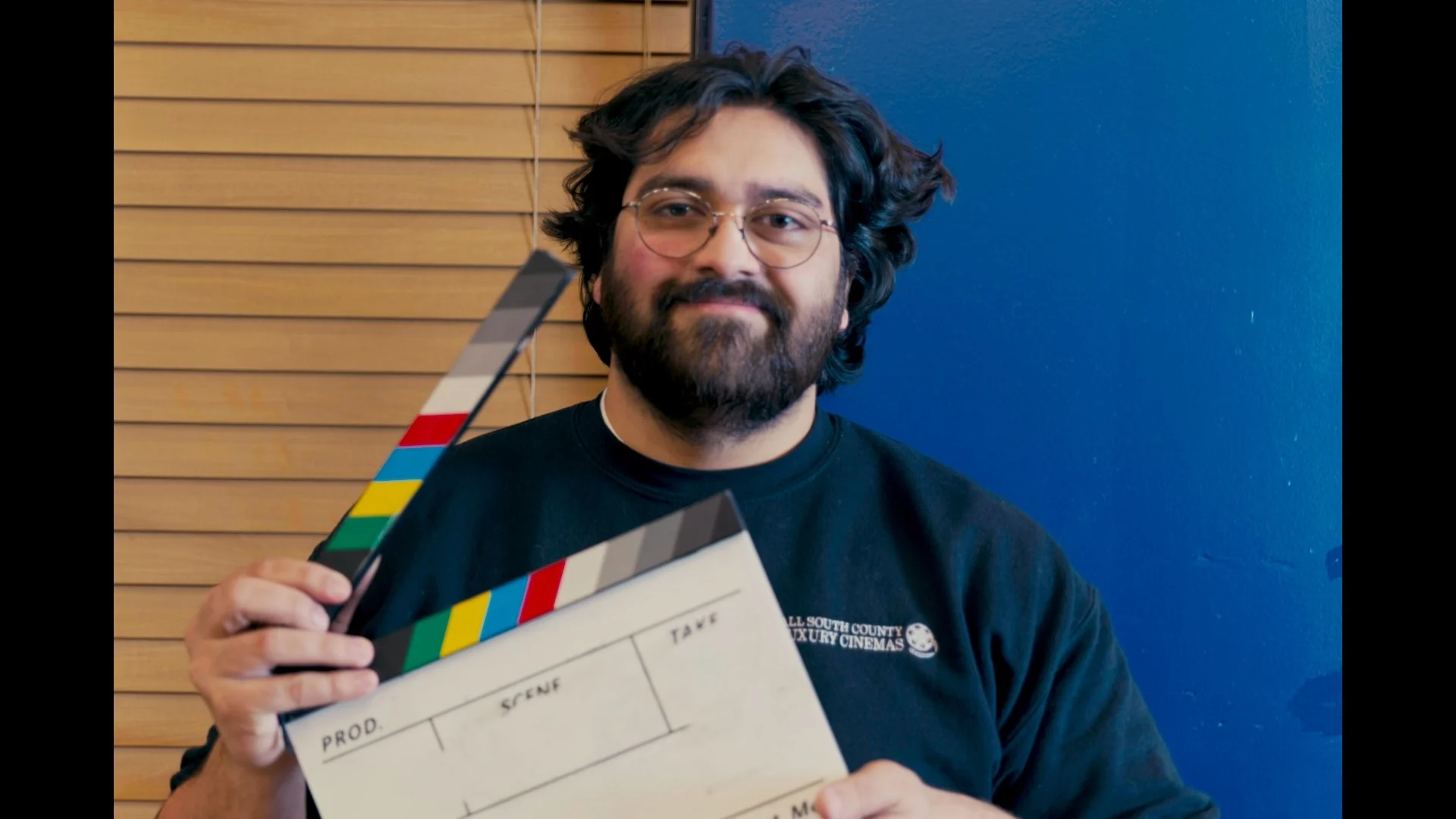 A man with glasses, a beard, and curly hair smiling while wearing a black Spider-Man t-shirt featuring Miles Morales and Spider-Man art.