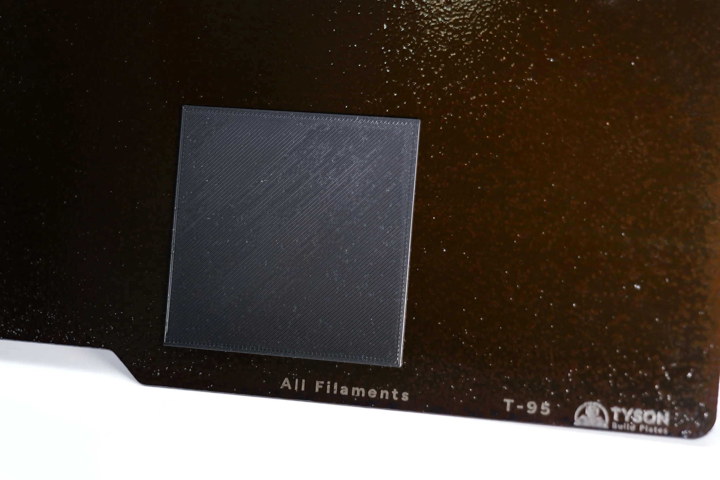 Close-up of a black panel with a textured surface, labeled "All Filaments" and "T-95," with a small part of the logo and text "TYSON Build Plates" visible at the bottom right corner.