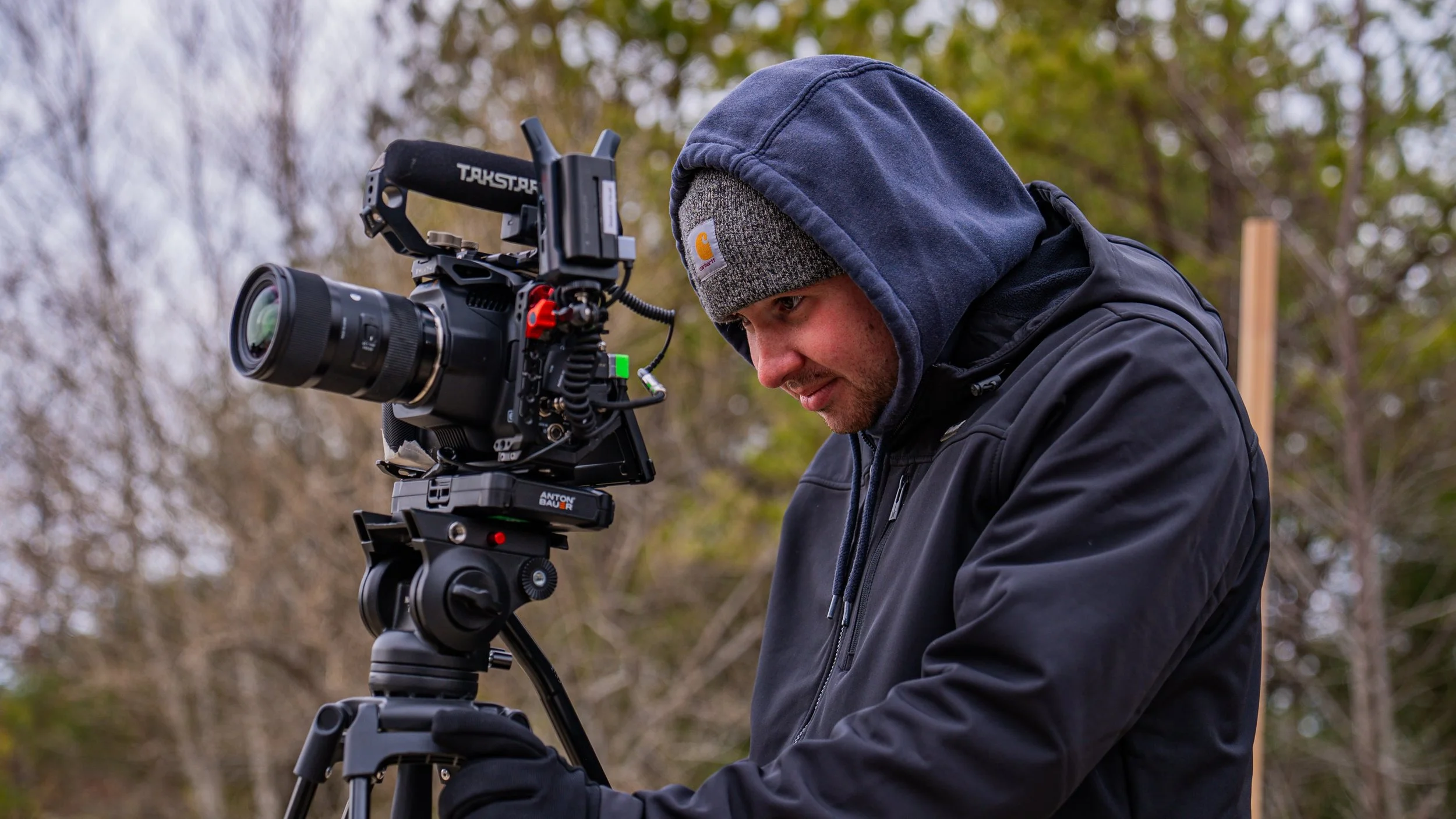 A person wearing a gray Carhartt beanie and a navy hoodie using a professional video camera mounted on a tripod outdoors during the day.