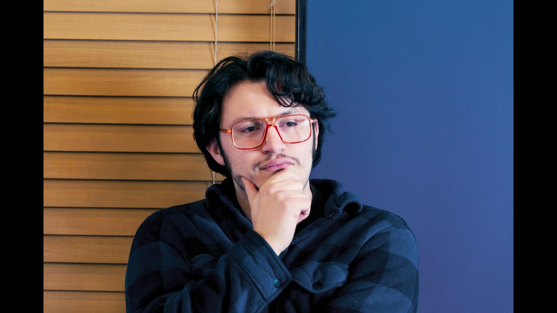 A person with dark hair and glasses holding a snake indoors near a window with blinds.