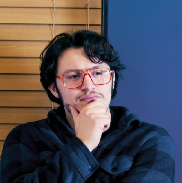 A young man with dark, wavy hair and glasses holding a snake in his hands indoors near a window with closed blinds.