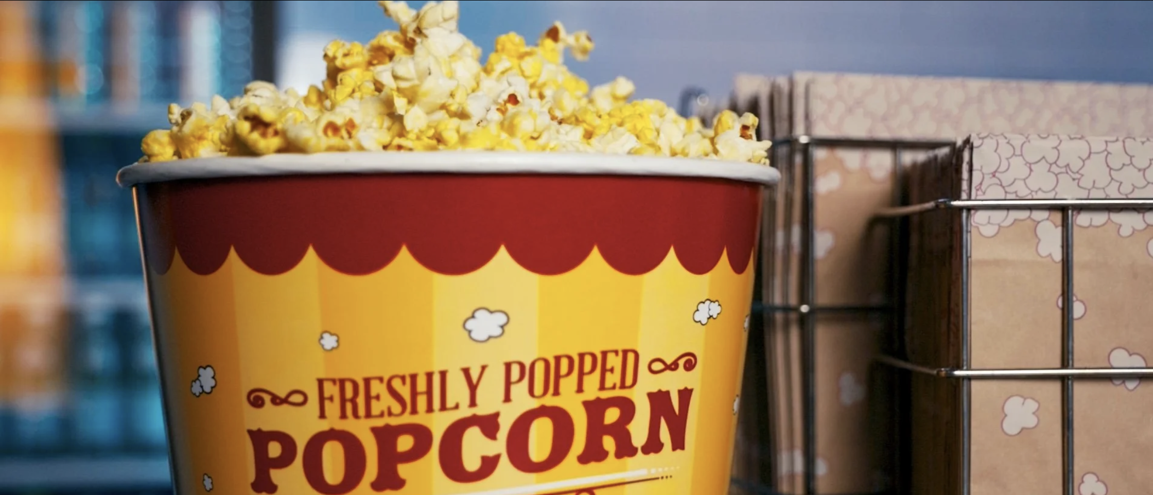 A red and yellow container of freshly popped popcorn with a cartoon-style design, placed on a shelf next to a wire basket holding boxed snacks.