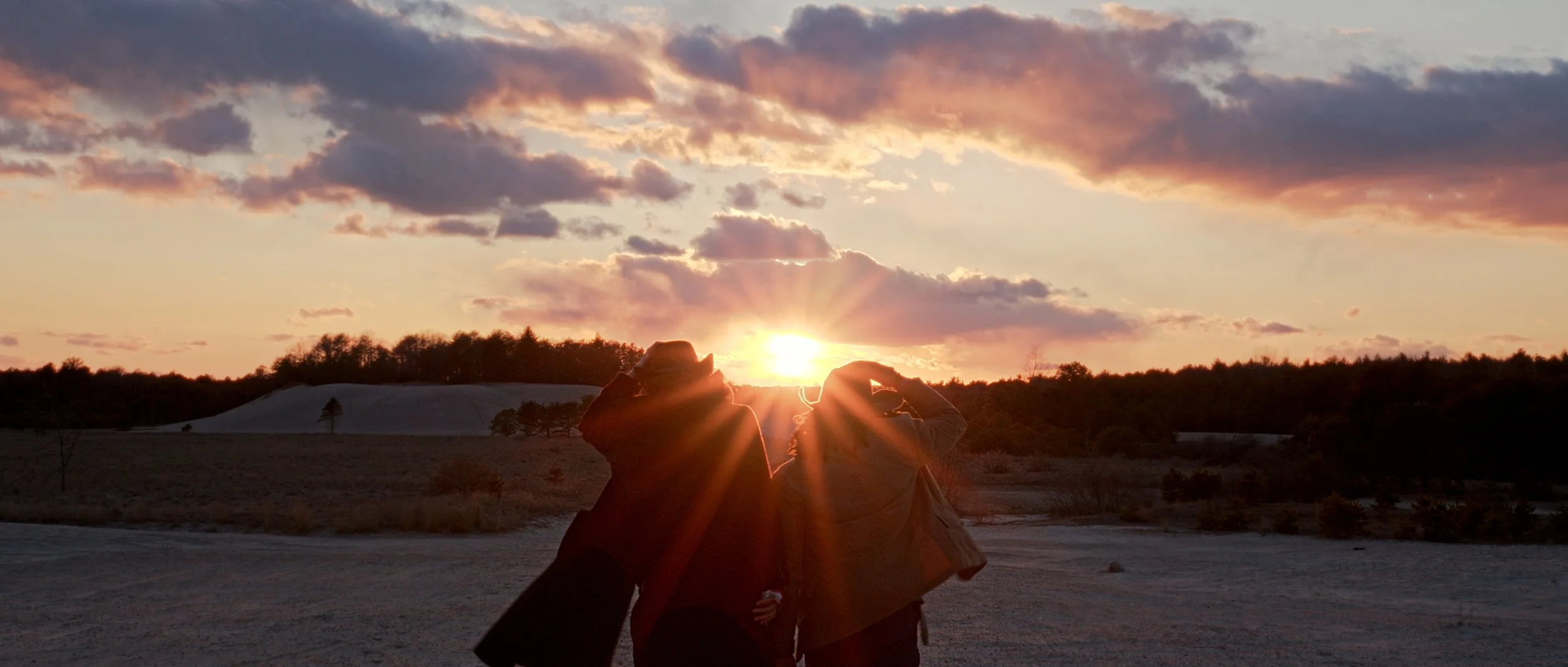 Two people standing outdoors at sunset, silhouetted against the sky with clouds, with a tree-lined horizon in the background.