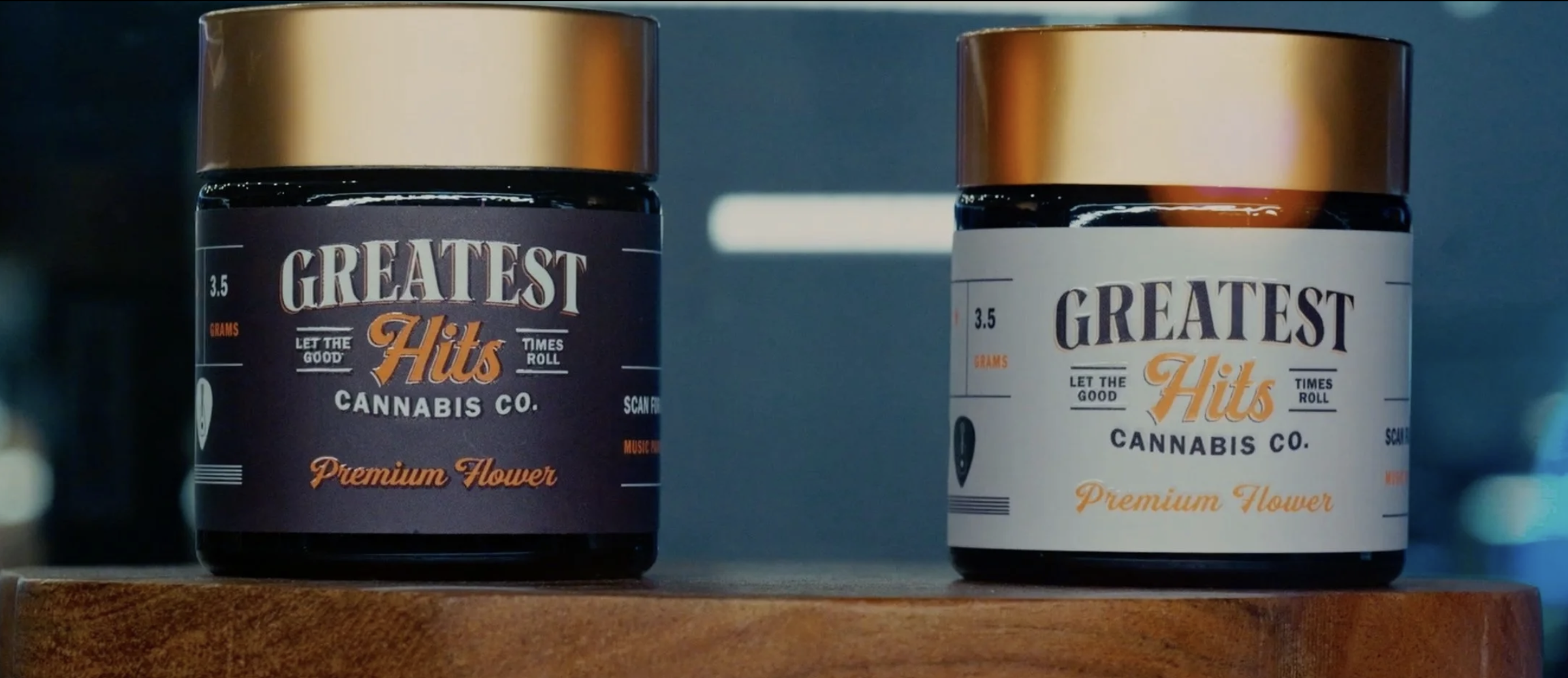 Two containers of cannabis products labeled "Greatest Hits Cannabis Co. Premium Flower" with one in black and the other in white packaging, sitting on a wooden surface.
