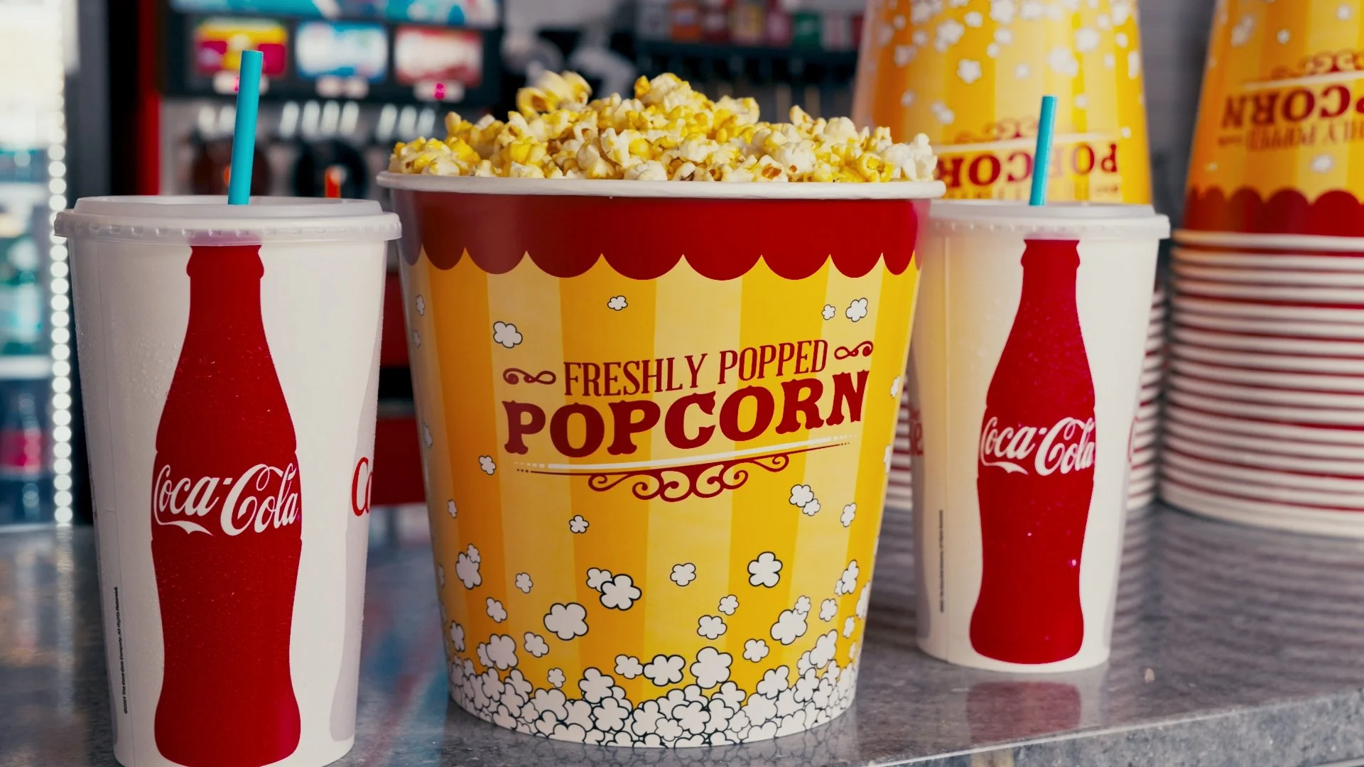 A large yellow striped bucket of freshly popped popcorn with the label 'Freshly Popped Popcorn', two cups of Coca-Cola with blue straws on either side, and additional cups and popcorn buckets stacked in the background.