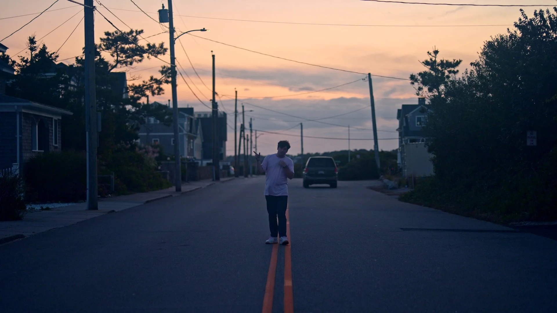 A young man walking on a deserted street at sunset, with utility poles and houses lining the road, and a car parked in the distance.