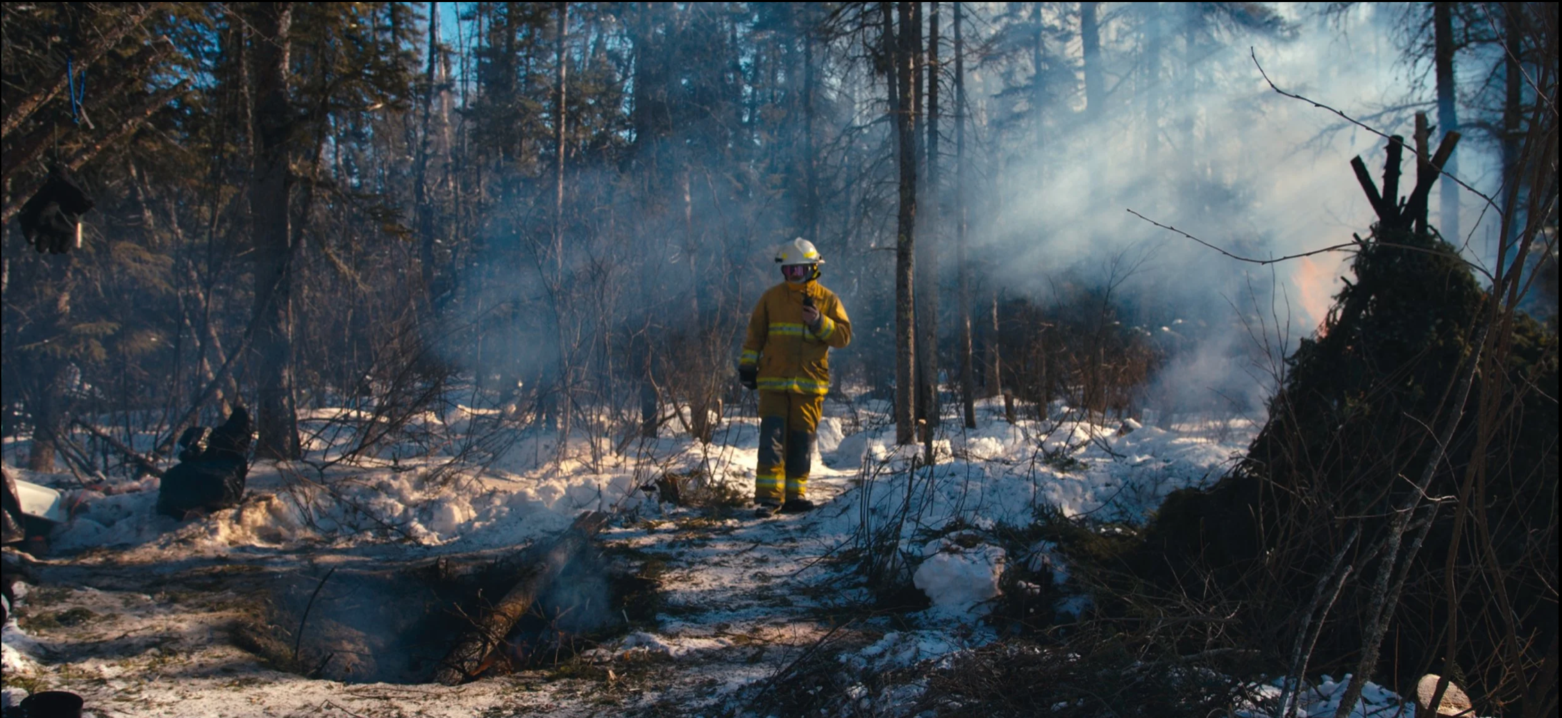 Firefighter in yellow uniform and helmet walking through a snowy forest with smoke and burnt trees, indicating a recent fire.