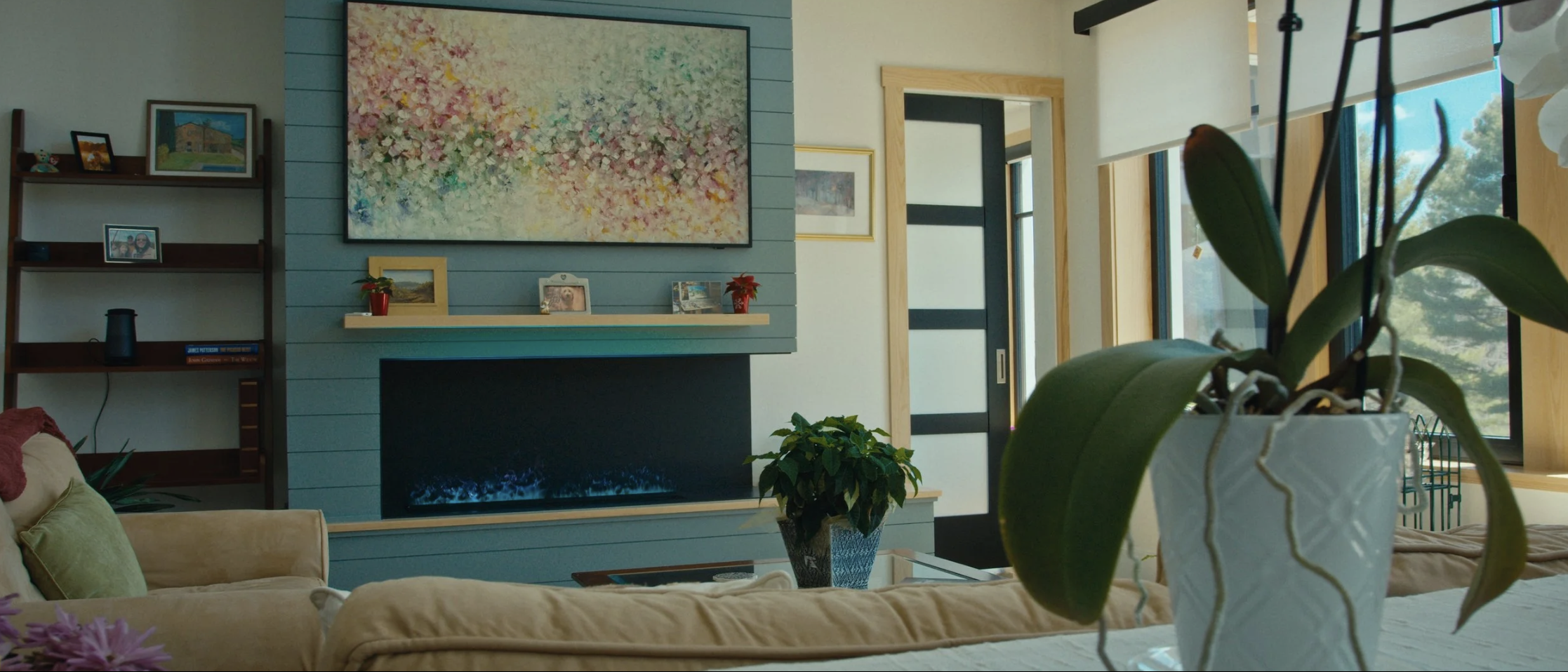 A living room with a beige sofa, a green cushion, a white table with a potted orchid, a fireplace, and a wall with framed pictures and a large colorful abstract painting. Sunlight comes through the windows, revealing trees outside.