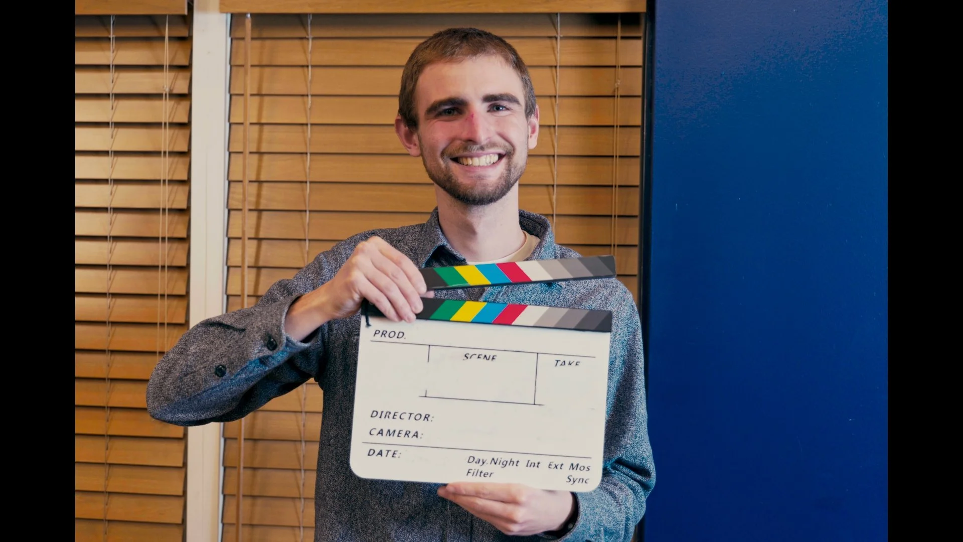 A young man with a beard and short hair sitting on a stone wall by the water, smiling, during sunset with a scenic view of the sea and distant islands.