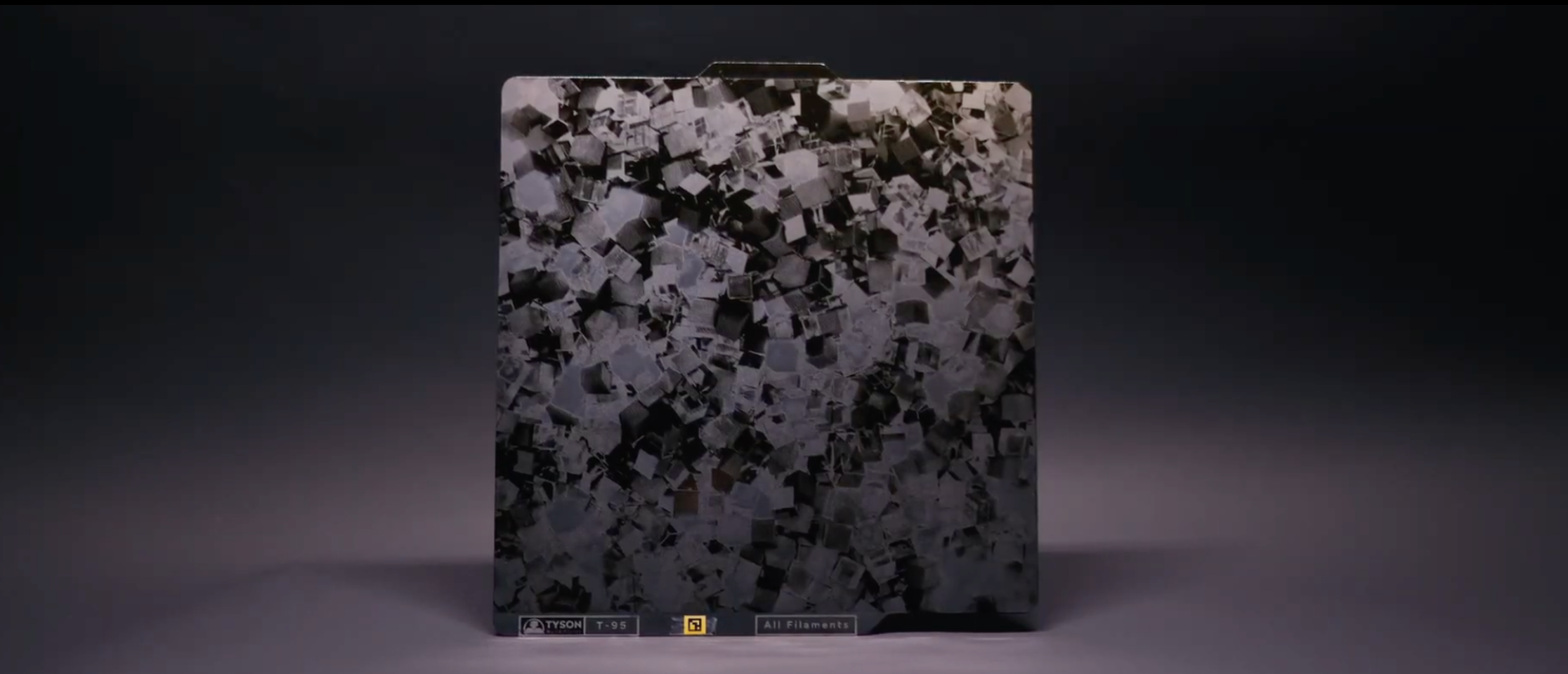 A display case with small, cube-shaped samples of materials or objects, with a dark background. The case is labeled with the Tyson logo, model number T-95, and a sign indicating 'All Filaments'.