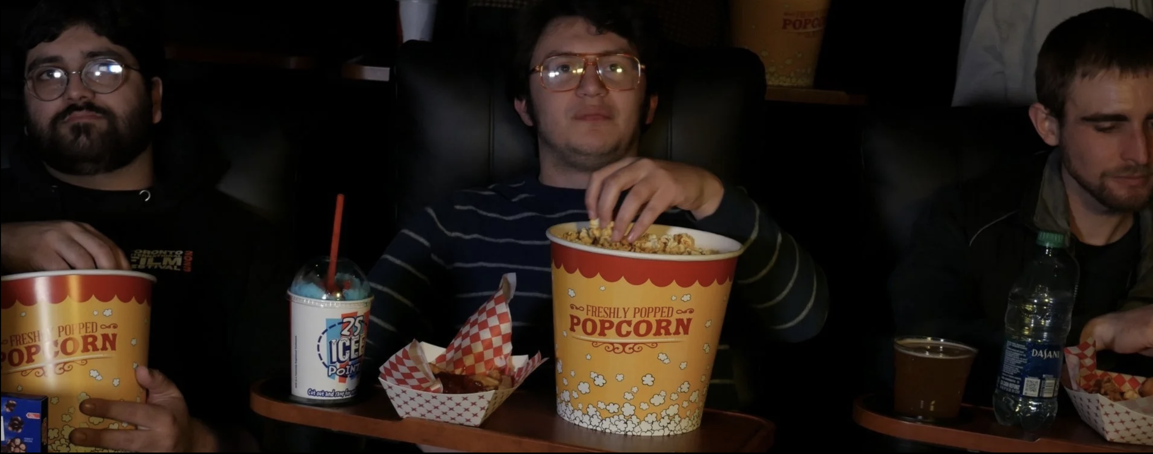 Three young men sitting at a table in a movie theater, eating popcorn and drinks, with one of them wearing glasses and striped shirt, surrounded by movie snacks.