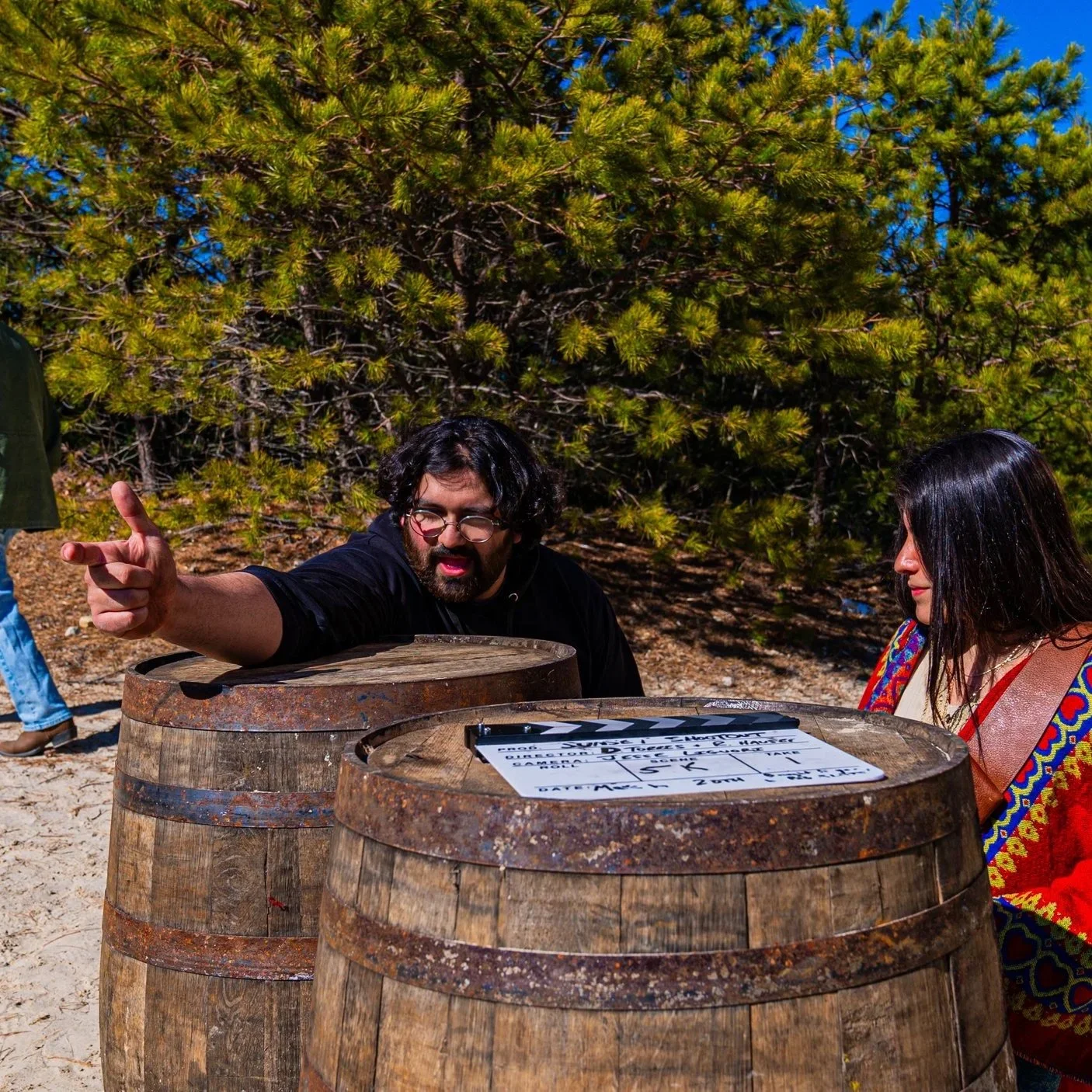 Two people talking behind two wooden barrels outdoors with green trees and blue sky in the background; one man with glasses and dark curly hair pointing, and a woman with long black hair wearing a colorful shawl, both focused on a piece of paper on one of the barrels.