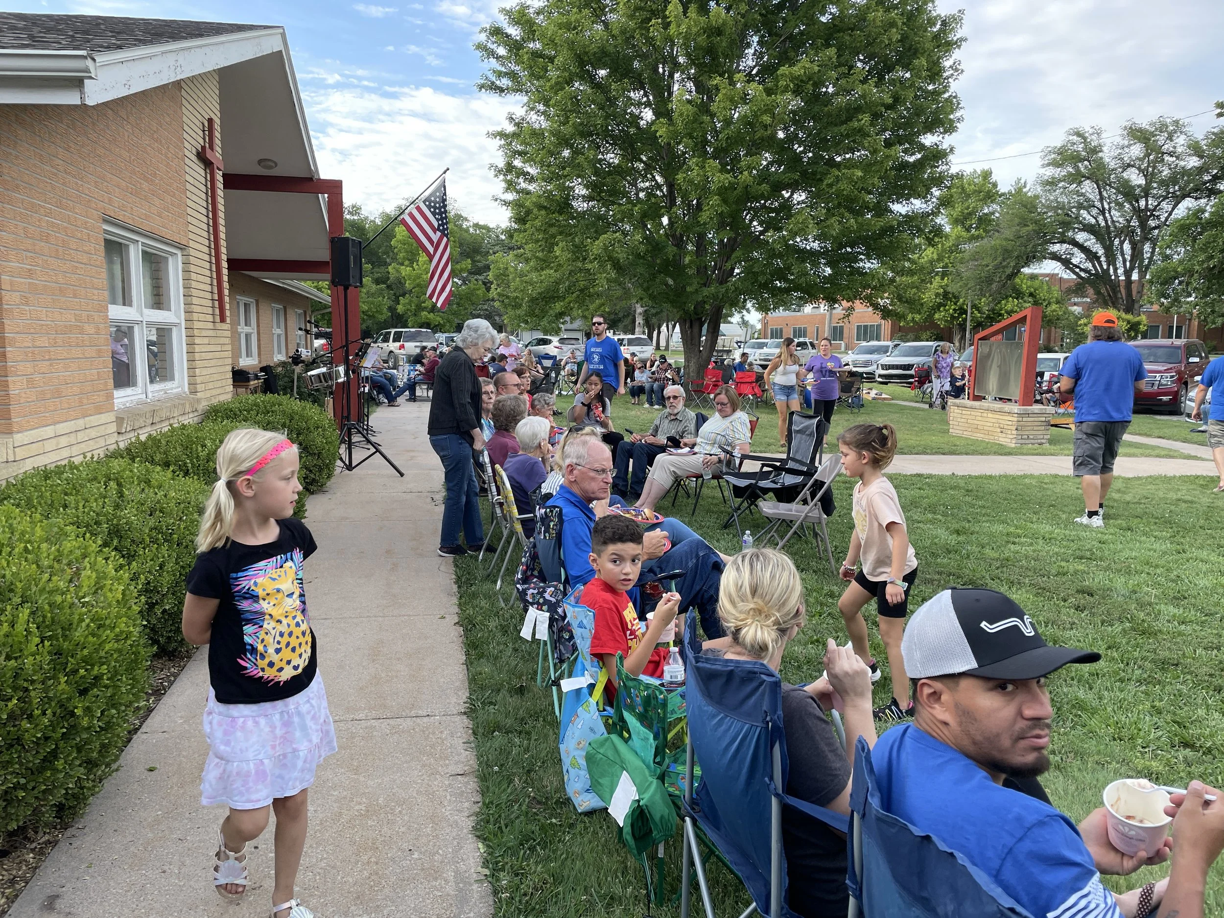 People sitting and standing outside a building, some children playing, with American flags seen on the building, a large green tree, and parked cars in the background, indicating a community gathering or outdoor event.