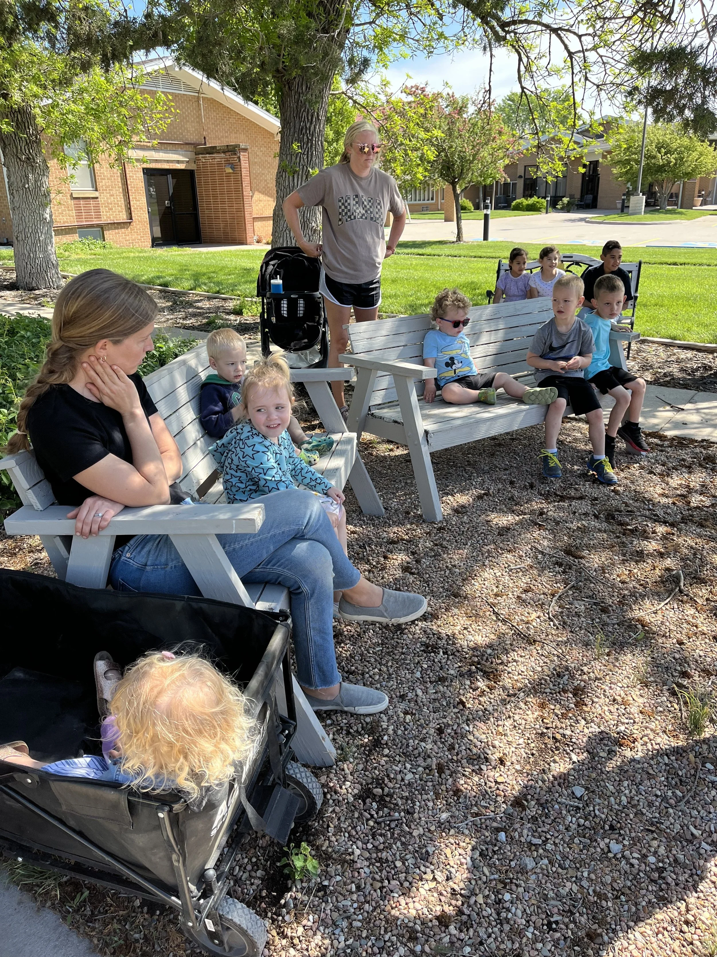 A group of children and two women sitting and standing on park benches under trees in a sunny park, with a stroller and a brick building in the background.
