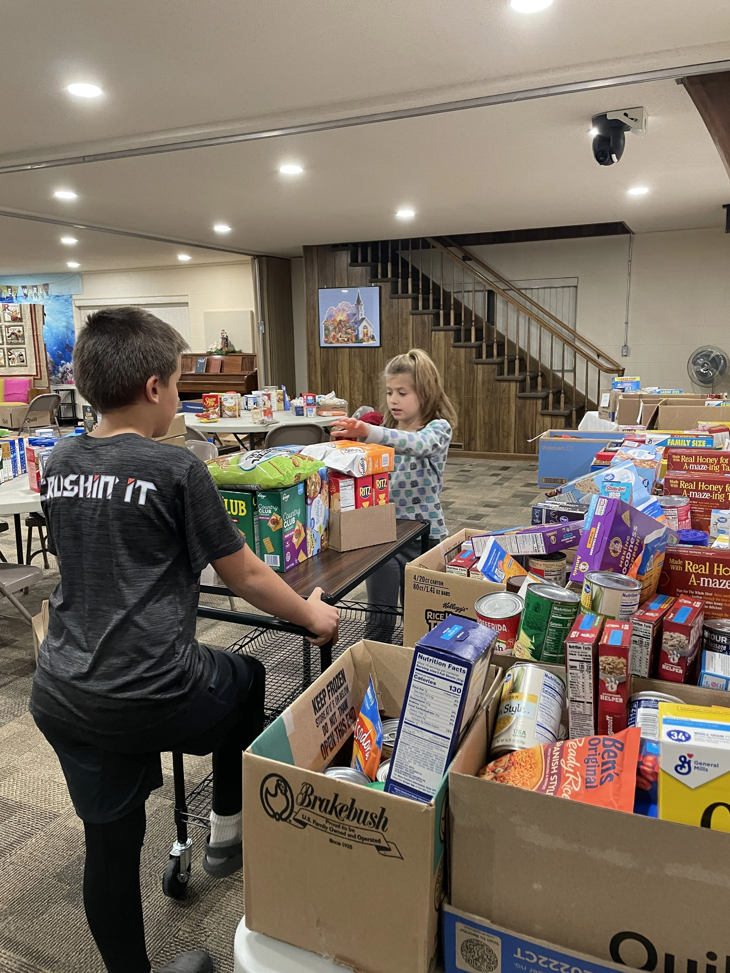 Two children, a boy and a girl, are organizing food items on tables and in boxes in a community center or similar setting for a food drive.