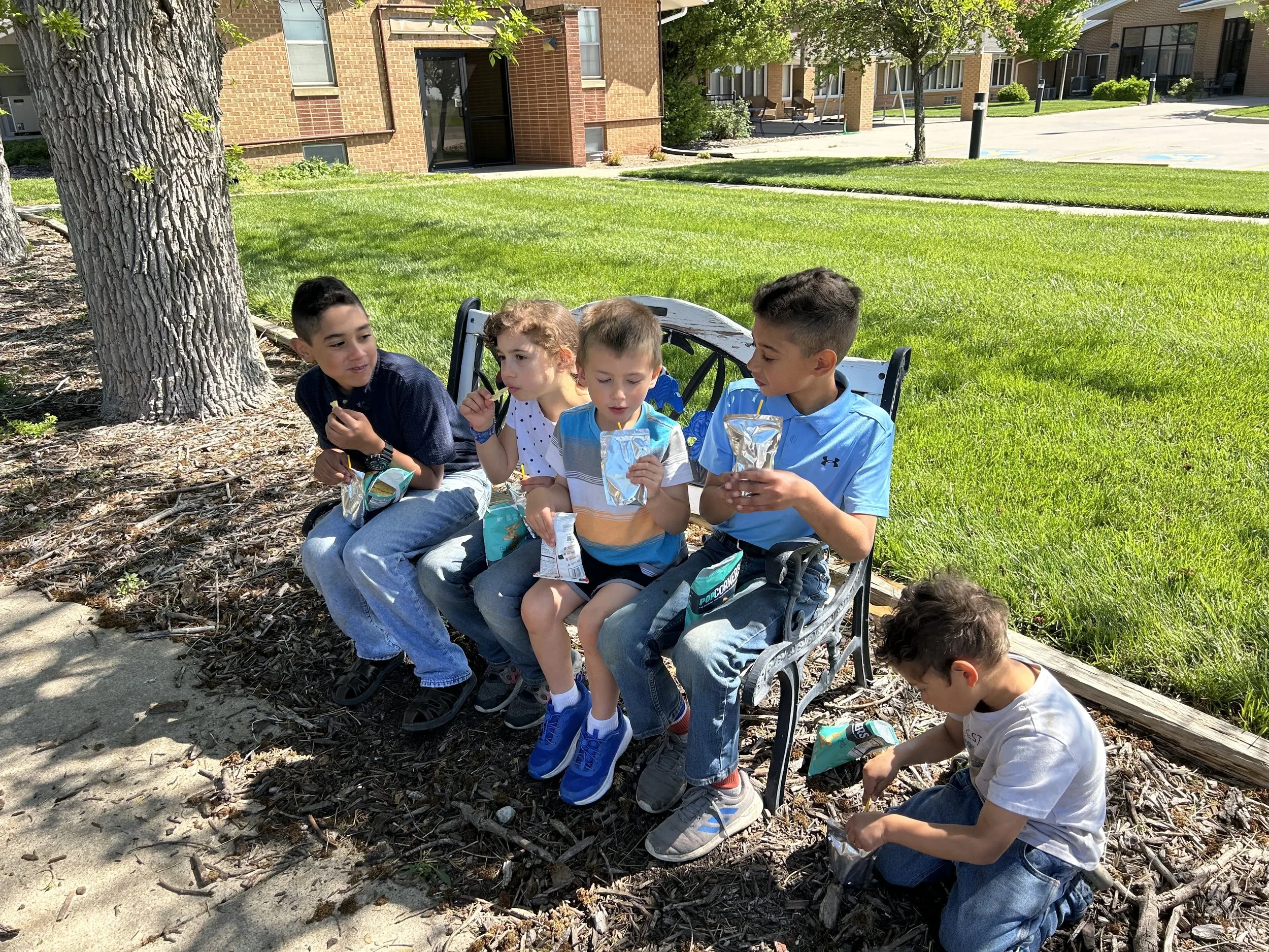 Five children sitting on a park bench and the ground, eating snacks on a sunny day, with green grass, trees, and apartment buildings in the background.