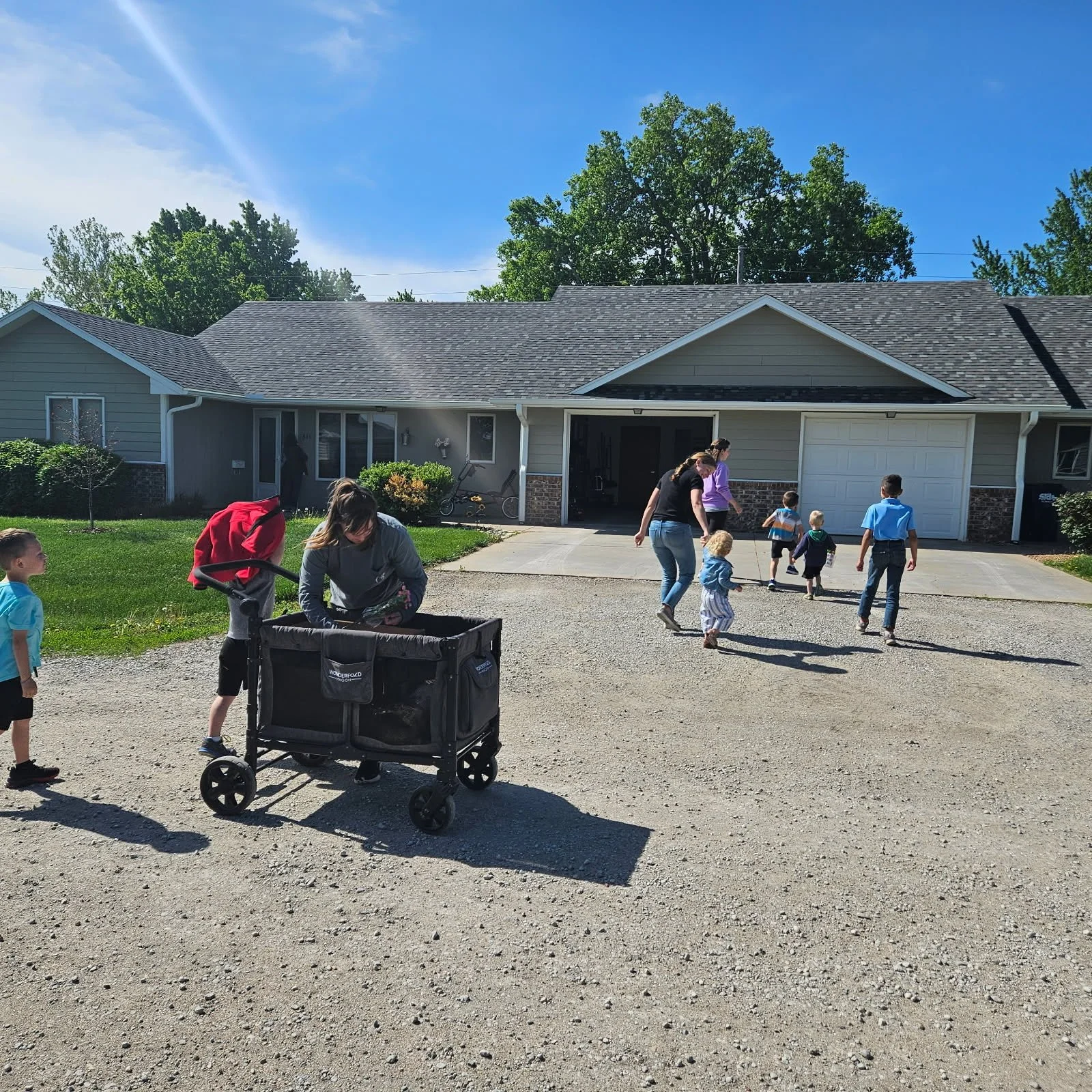 Children and adults playing and walking outside a house with a garage and driveway on a sunny day.