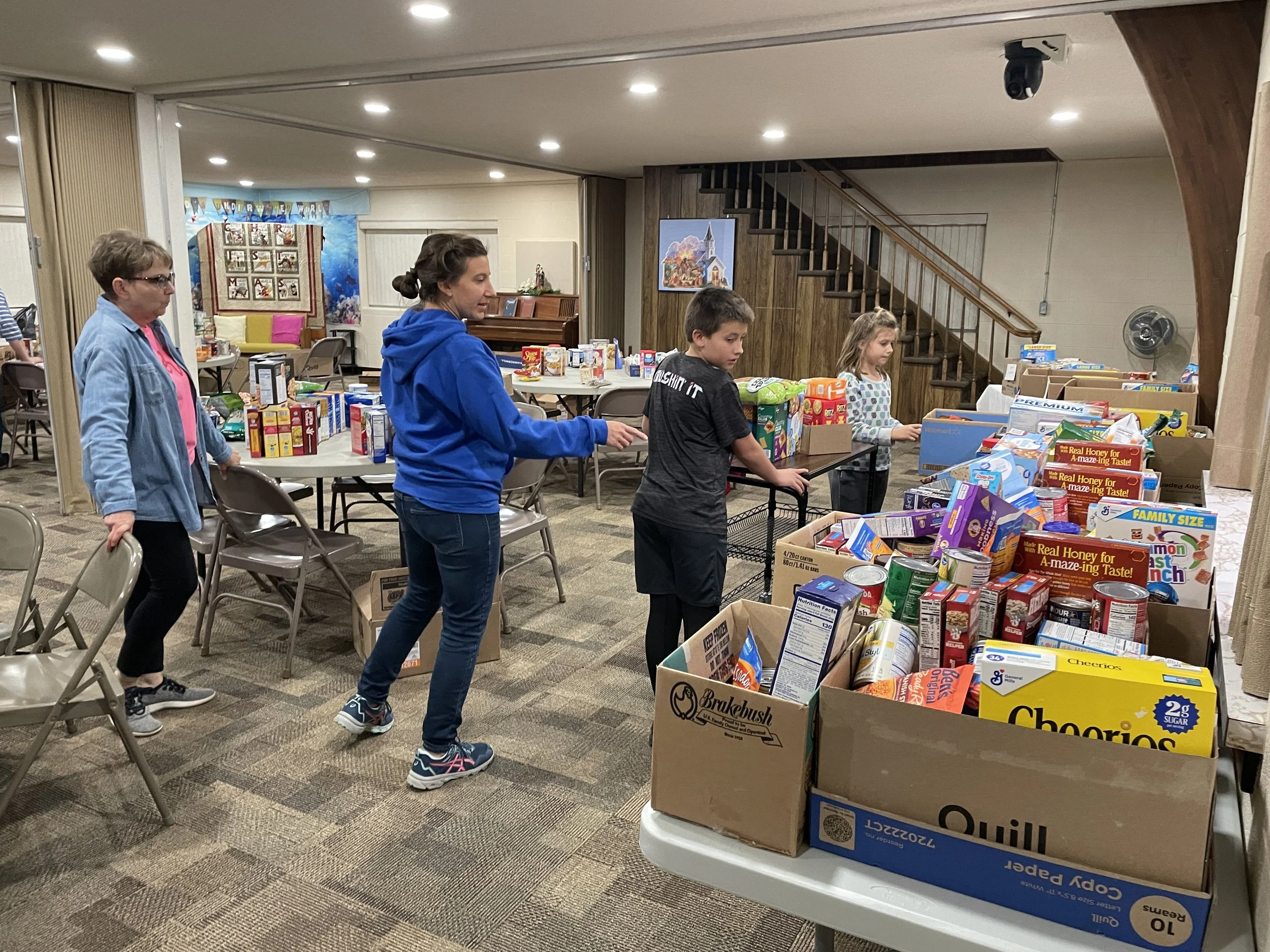 People organizing and packing food and supplies in a room, with boxes filled with various food items on tables.