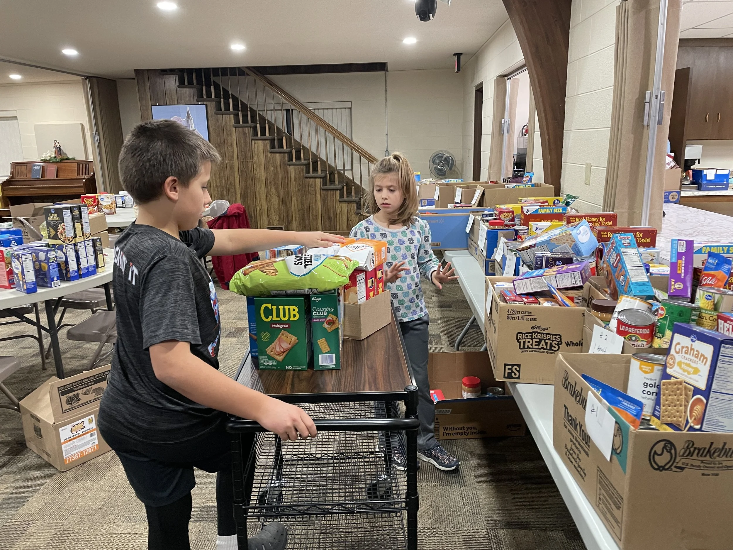 Two children, a boy and a girl, are shopping for food items in a room with tables and boxes of groceries. The boy is reaching for a bag of chips, while the girl looks at the food on the table.
