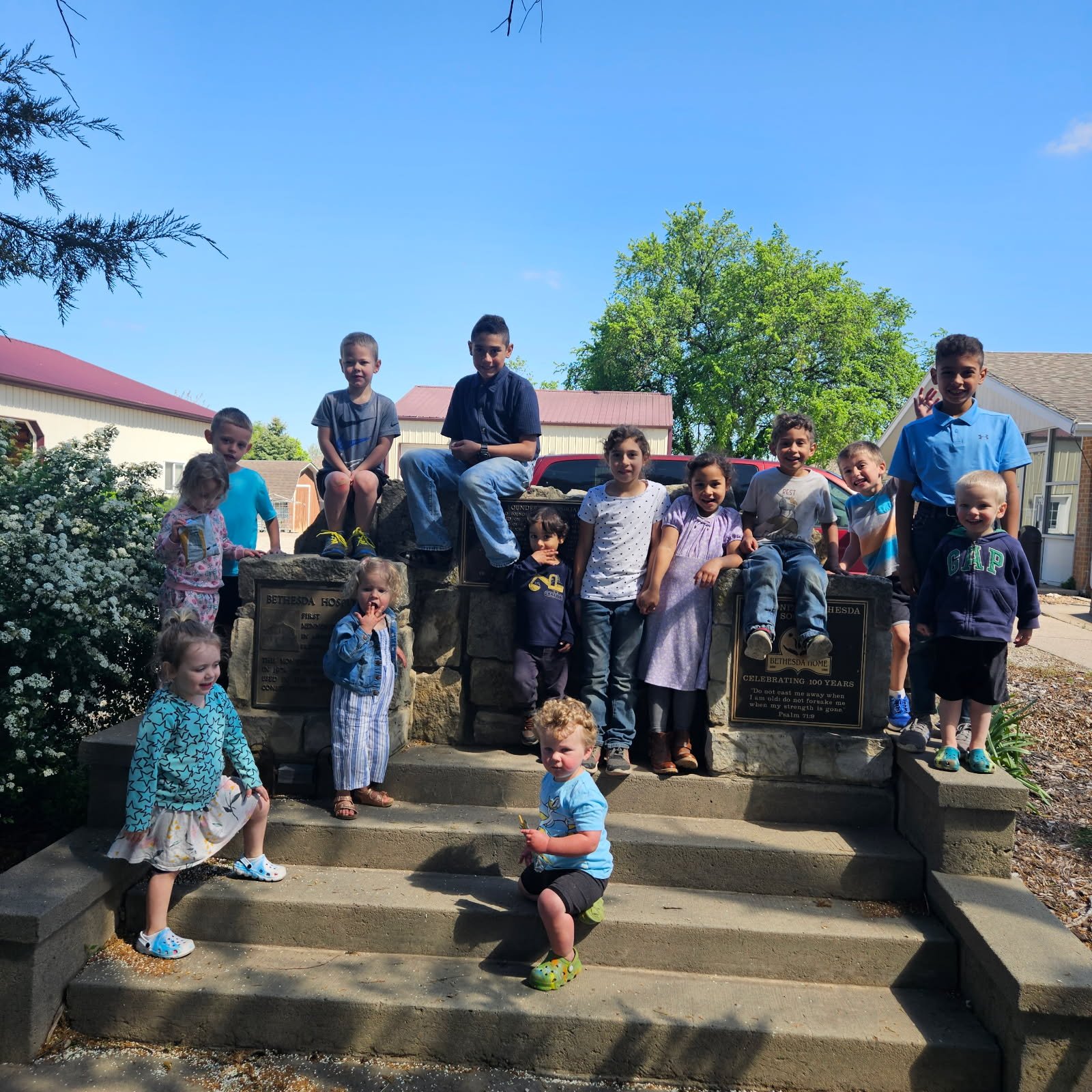 A group of children gathered around a stone monument outdoors on a sunny day, with trees and houses in the background.