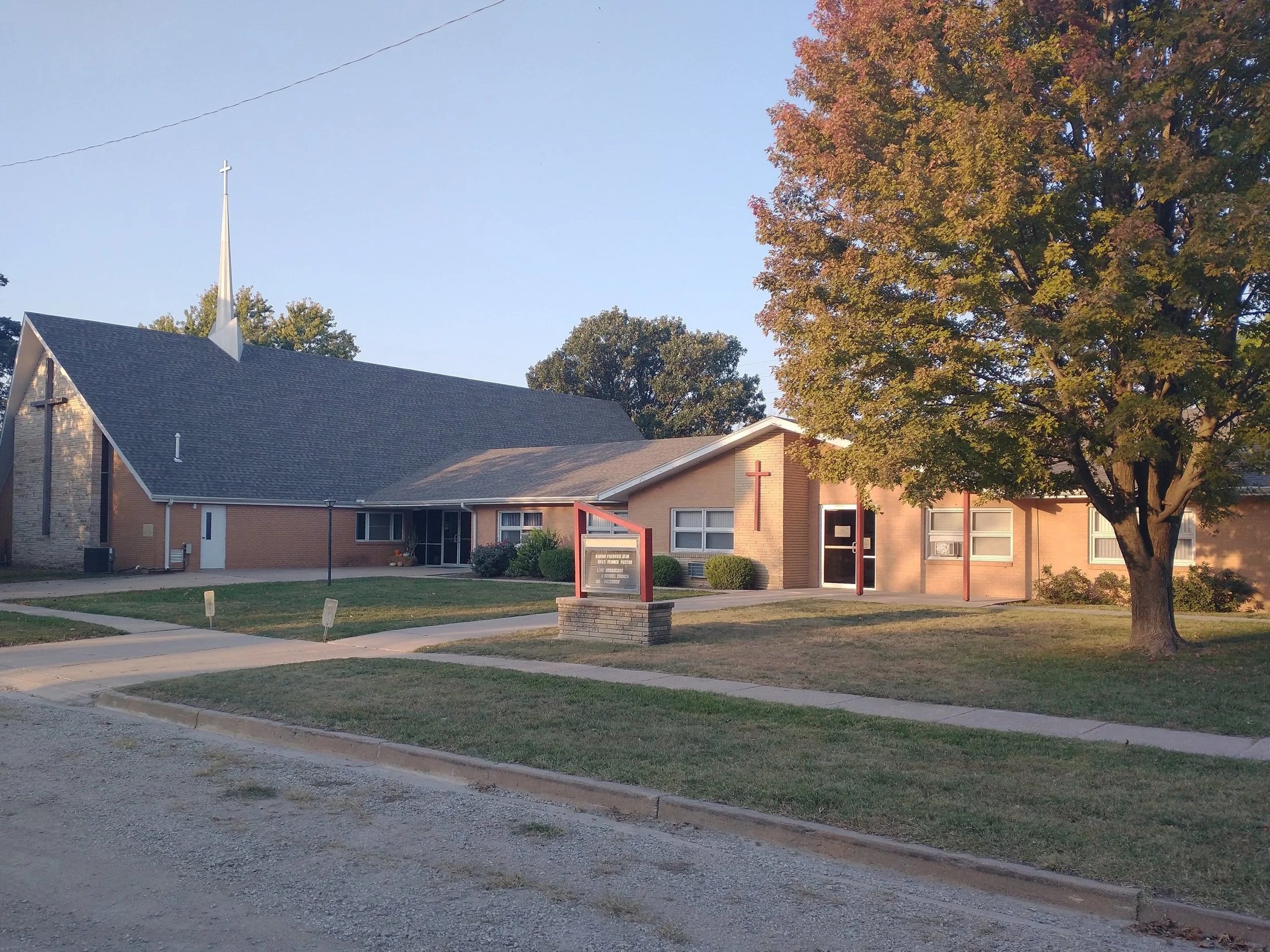 A church building with a brick exterior, a tall steeple with a cross at the top, a large tree in front, and a sign near the sidewalk, illuminated by sunlight in the late afternoon.