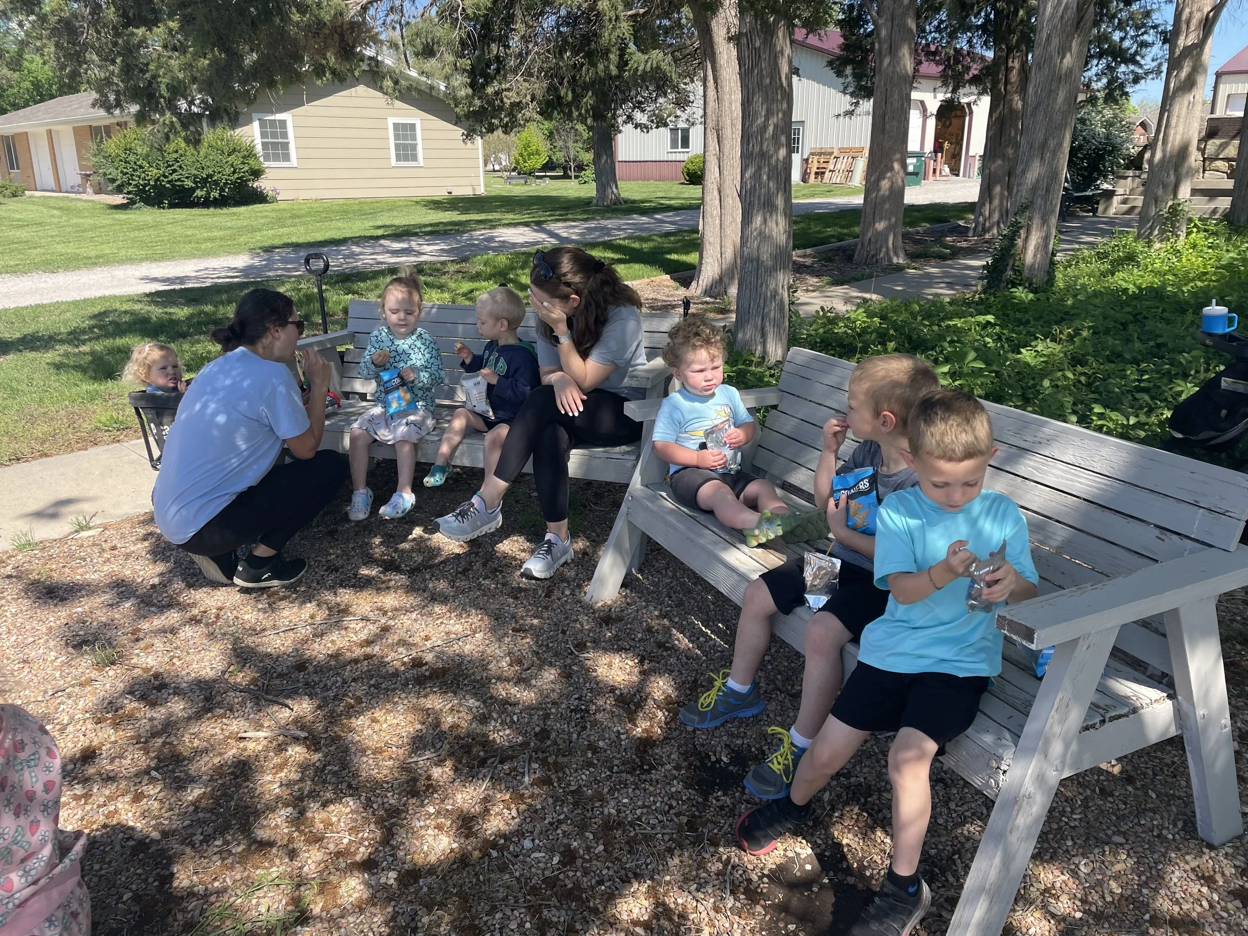 Group of children and two adults sitting on benches under a tree on a sunny day, enjoying snacks and drinks in a park.