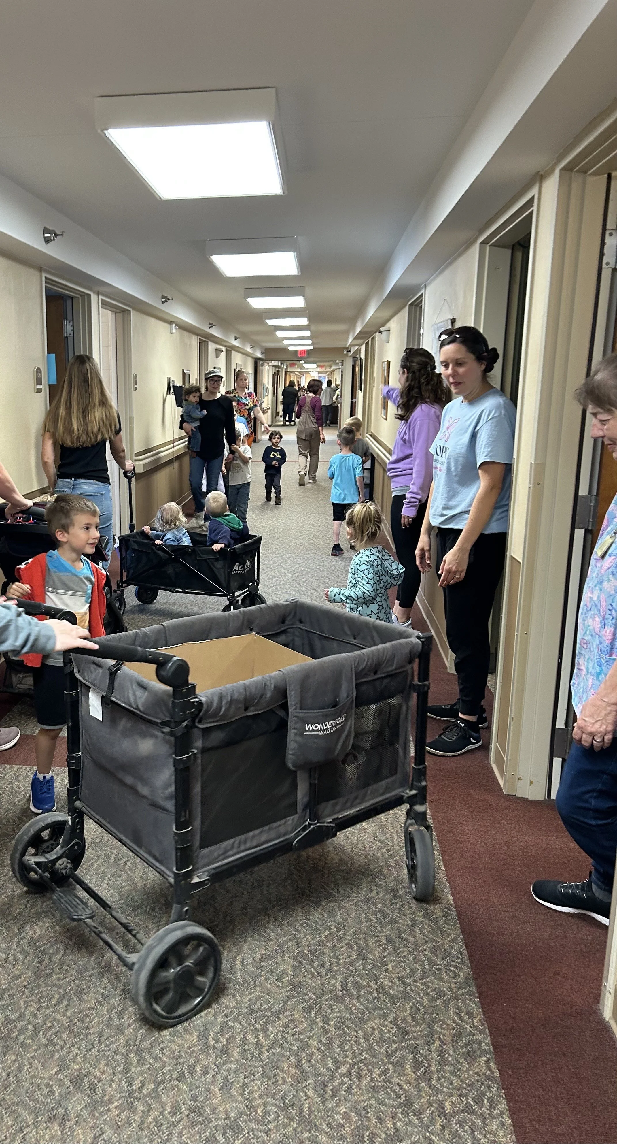 Children and adults gather in a hallway, some with strollers and carts, engaging in an activity or event.