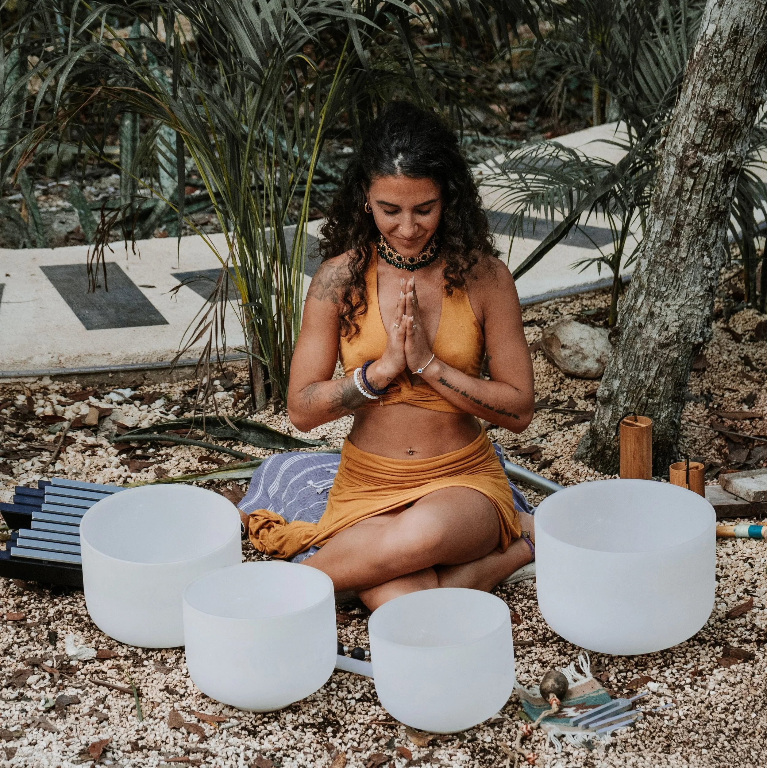 A woman with curly hair doing meditation outdoors surrounded by plants and trees, sitting cross-legged on a towel with singing bowls around her.