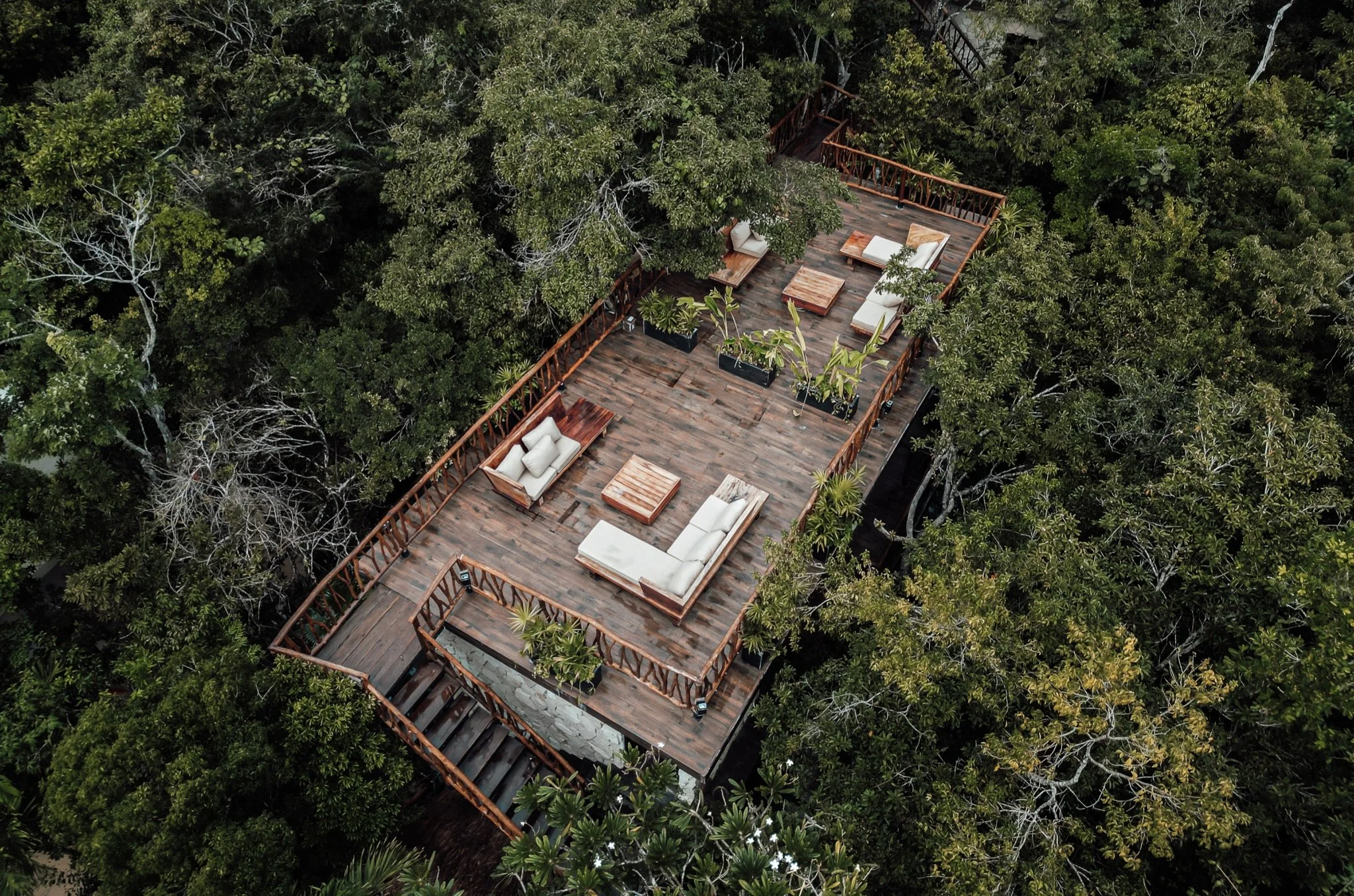 A wooden rooftop deck with outdoor furniture, including white cushioned sofas, chairs, and tables, surrounded by lush green trees.