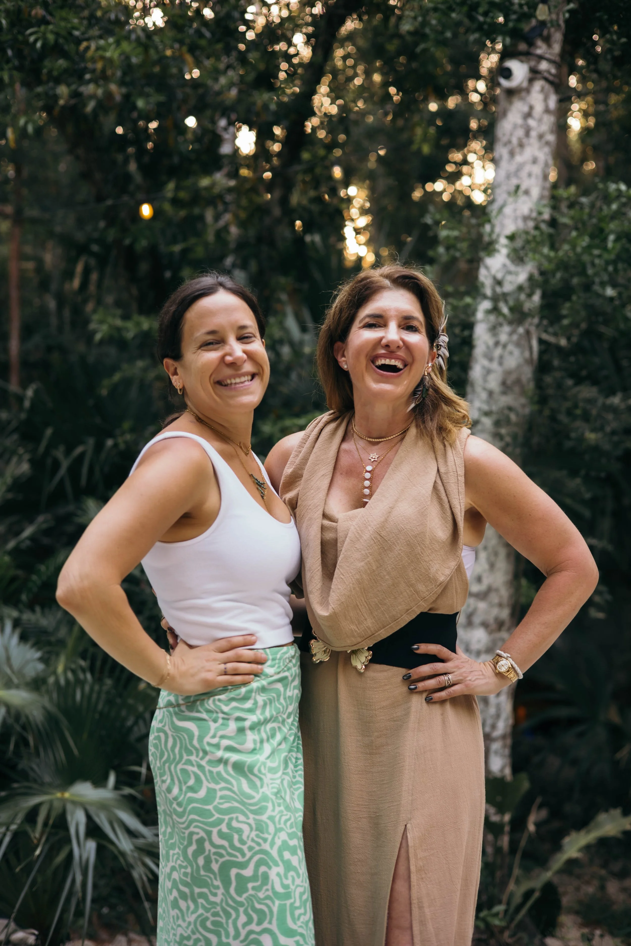 Two women smiling and posing outdoors in a forested area with trees and greenery.