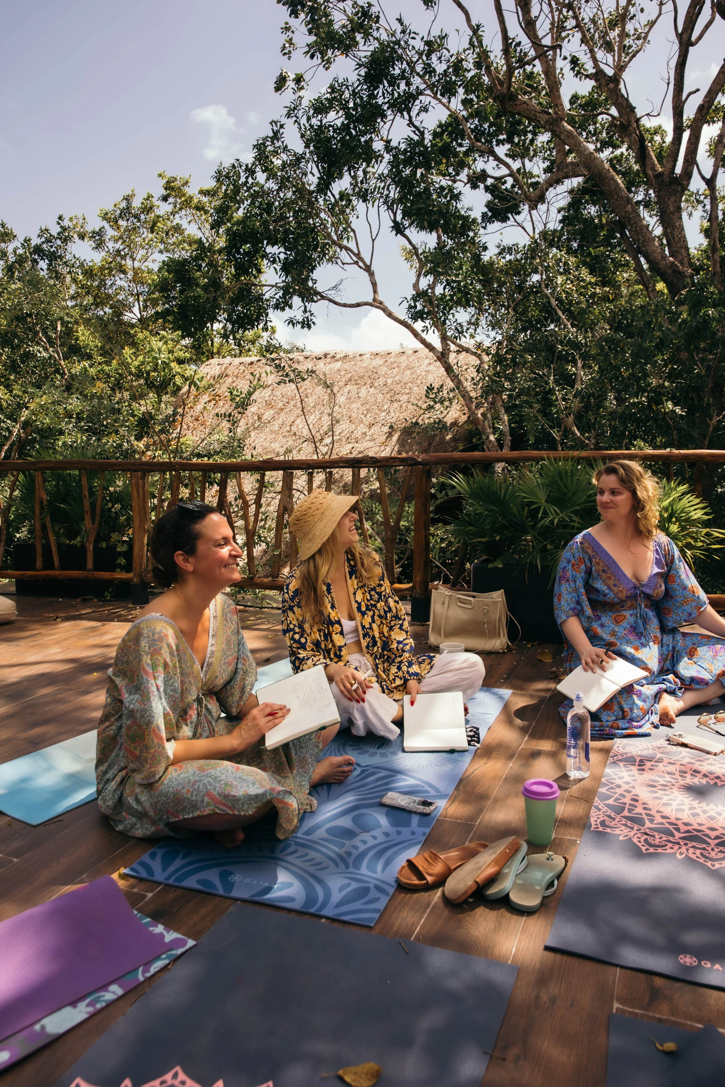 Three women participating in an outdoor yoga or meditation class on a wooden deck with yoga mats, surrounded by trees and greenery, one woman smiling while holding a notebook, another with a straw hat, and the third with curly hair.